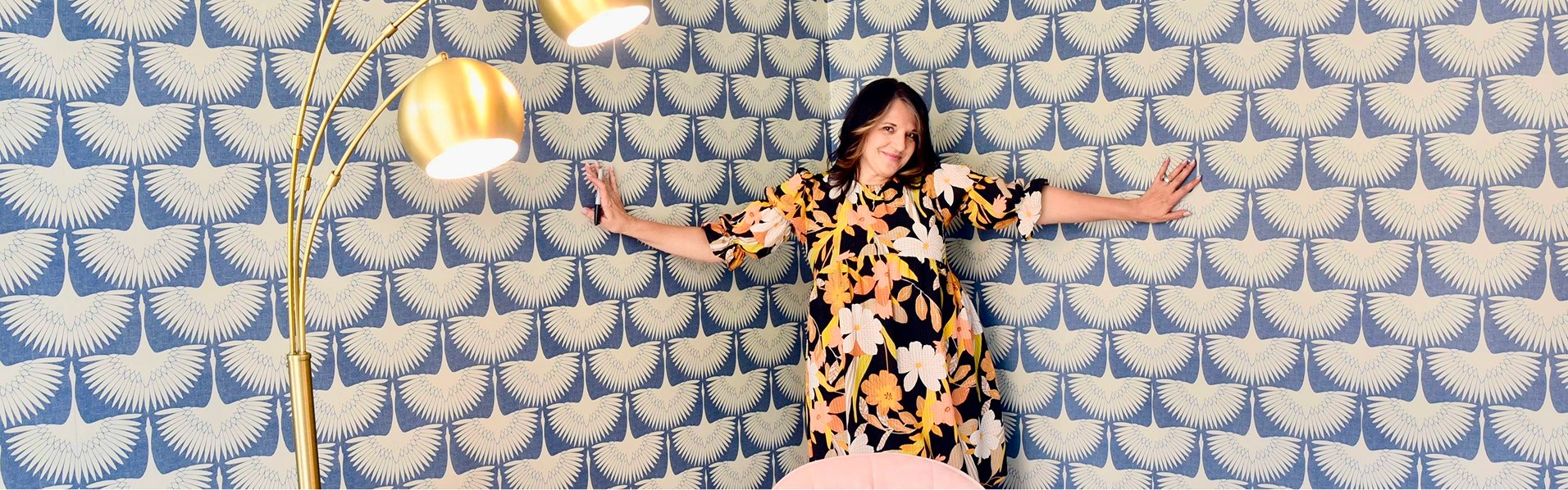 Smiling woman in floral dress with arms outstretched in stylish room of blue peacock wallpaper, gold lamps, and pink pouf.