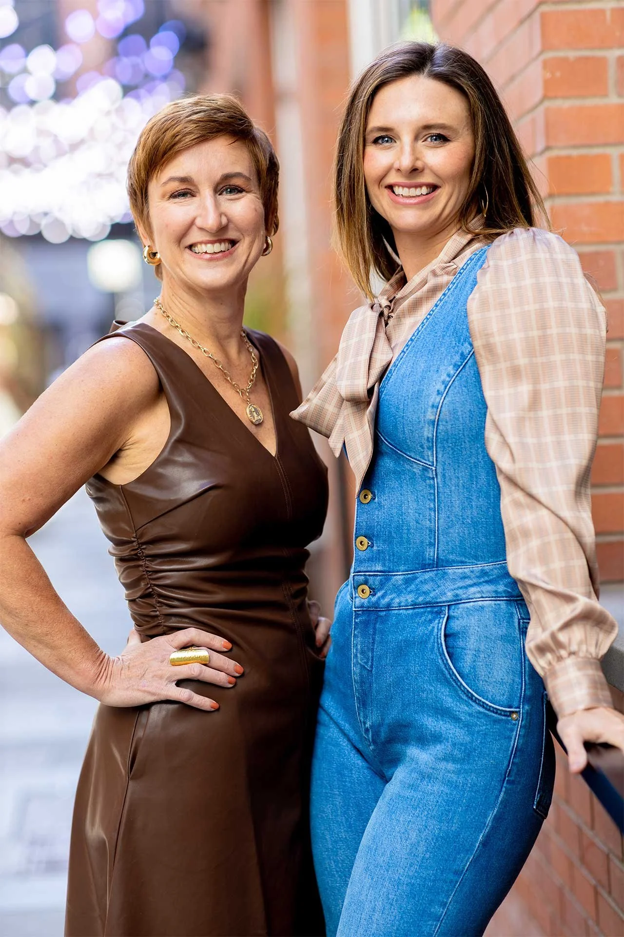 Portrait of Jen and Katie, staging home services professionals, posing confidently outdoors with urban brick backdrop and string lights.