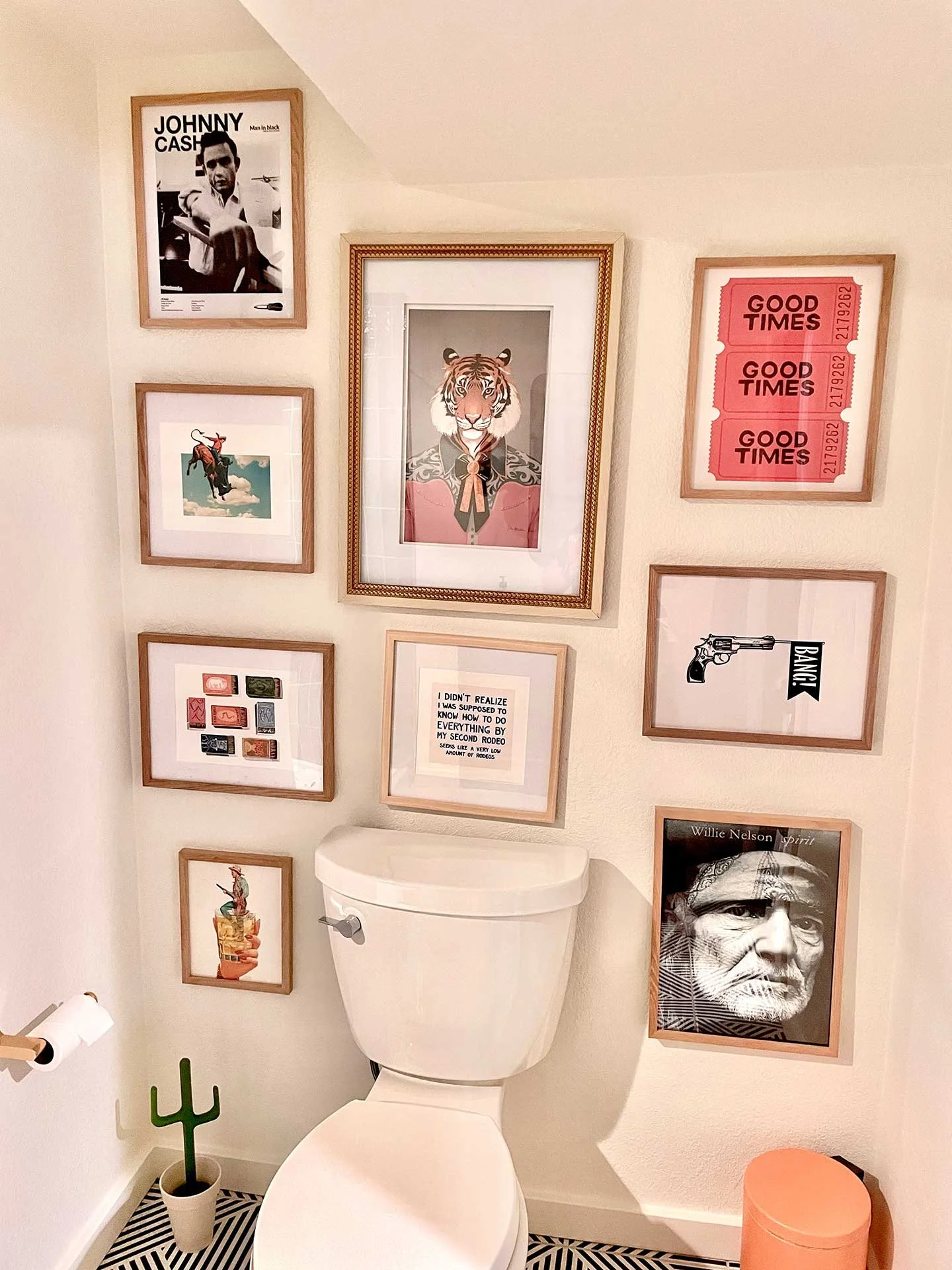 Eclectic gallery wall above the toilet with playful framed art, including tiger portrait, vintage tickets, typography, and Western-inspired prints in warm wood frames.