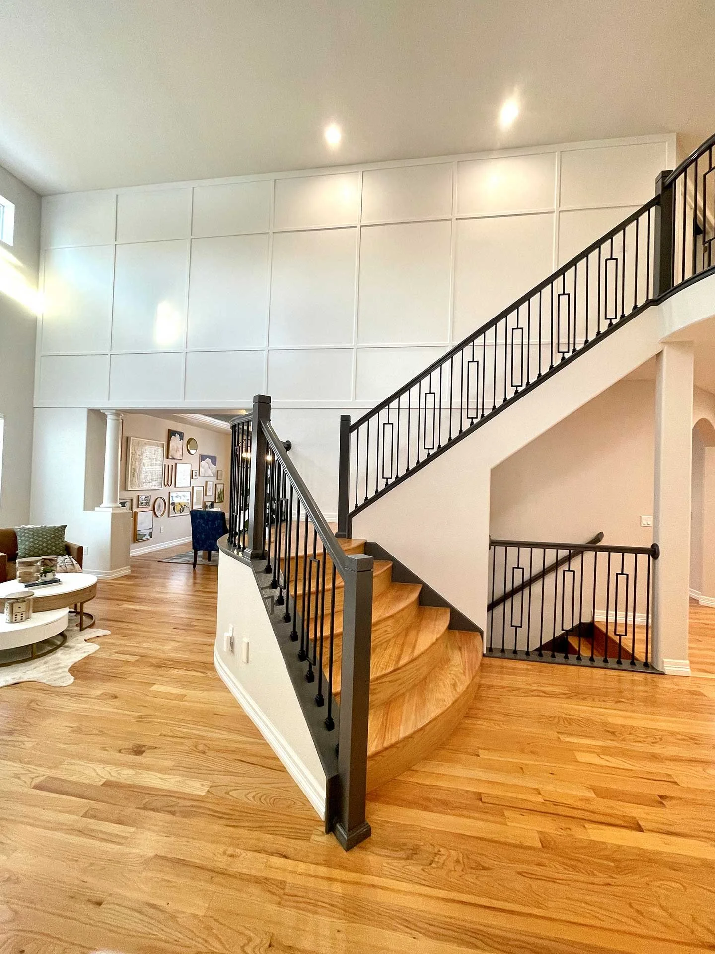 Open foyer with curved wood staircase, black metal railing, and large paneled accent wall, leading into a bright living area with warm wood flooring.