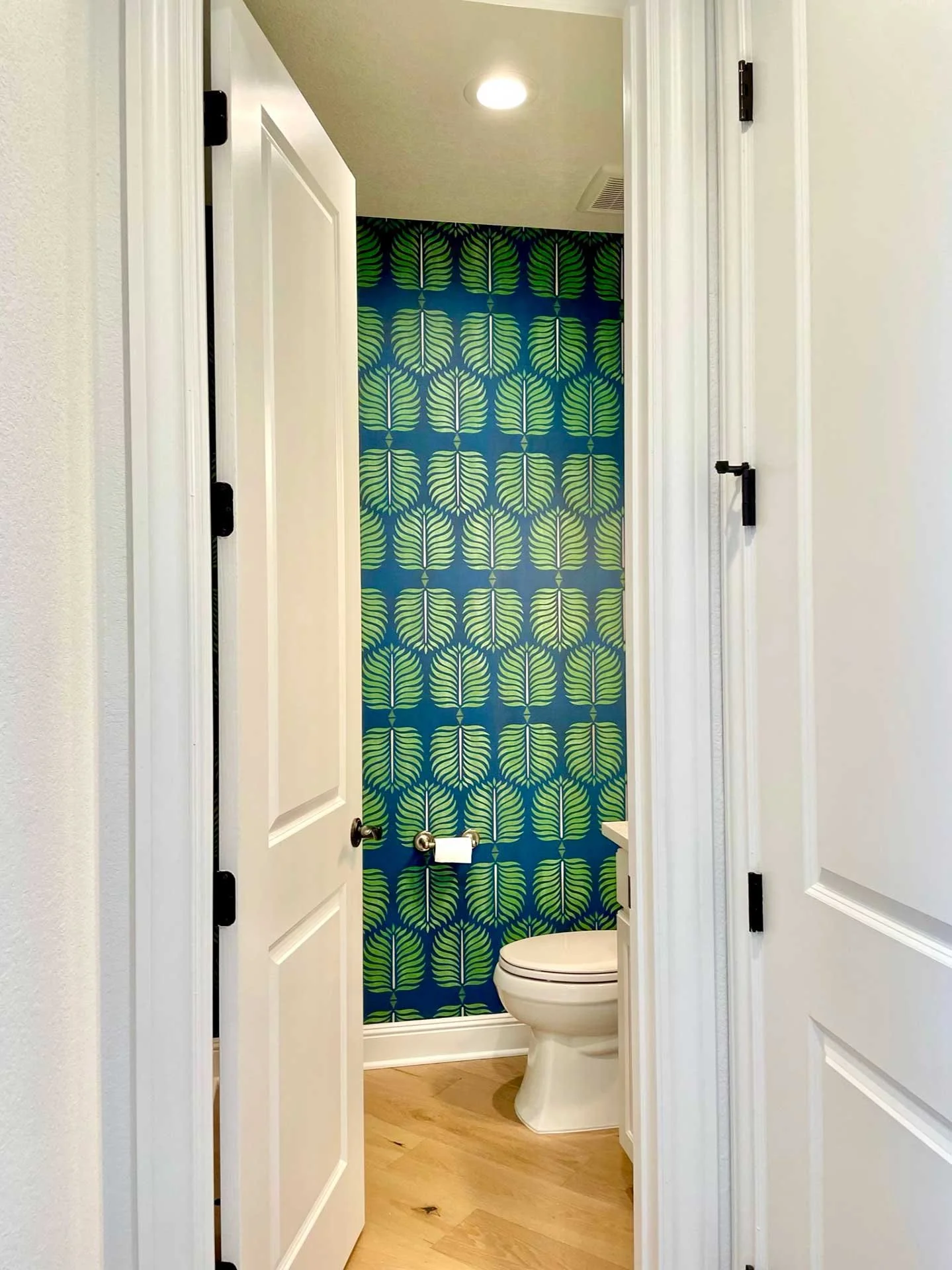 View into powder room with bold blue and green leaf-pattern wallpaper, white toilet, and light wood flooring framed by open doorway.
