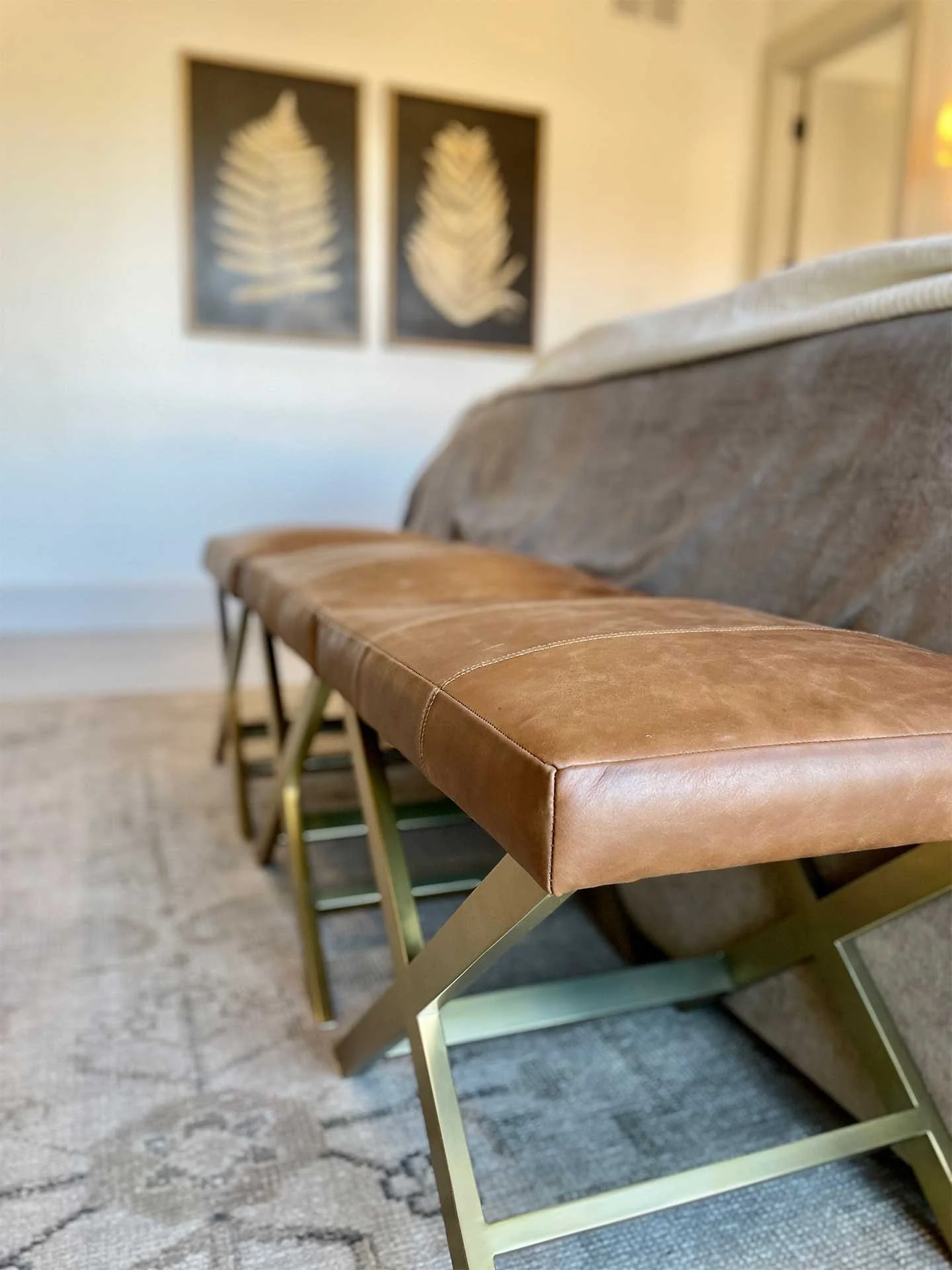 Luxurious tan leather bench on gold metal legs beside a bed with beige walls and fern art, after home staging services.