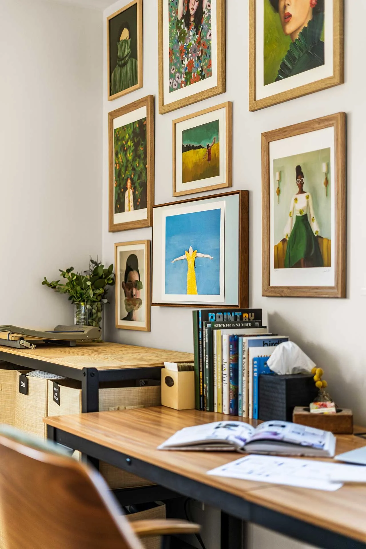 Home office nook with wood desk, open shelving, and gallery wall of colorful framed art, styled with books, greenery, and everyday essentials.