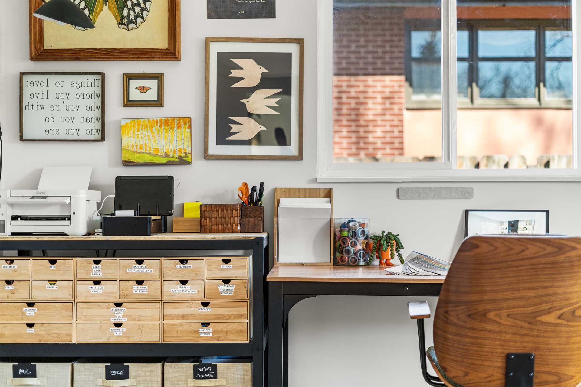 Desk setup with wall art of birds and landscape, wooden drawers, scissors, colorful mug, succulent, and window view