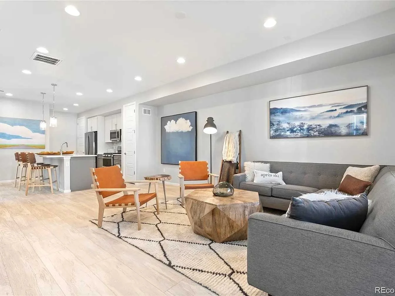 premium home staging of a den with white kitchen, bar stools, orange chairs, gray couch, and large blue artworks. 