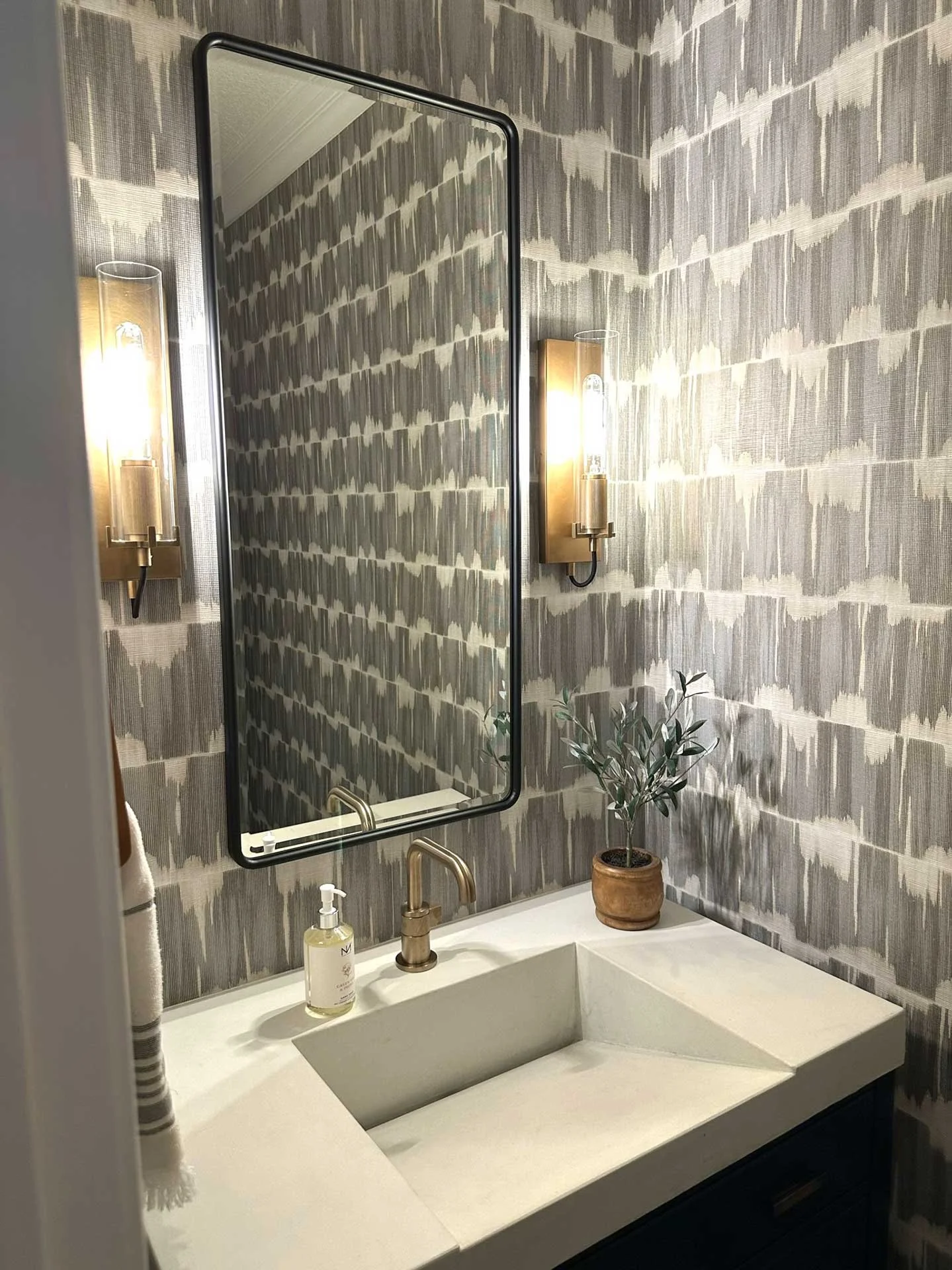 Modern powder room with gray patterned wallpaper, black-framed mirror, brass sconces, and integrated white sink with brass faucet.