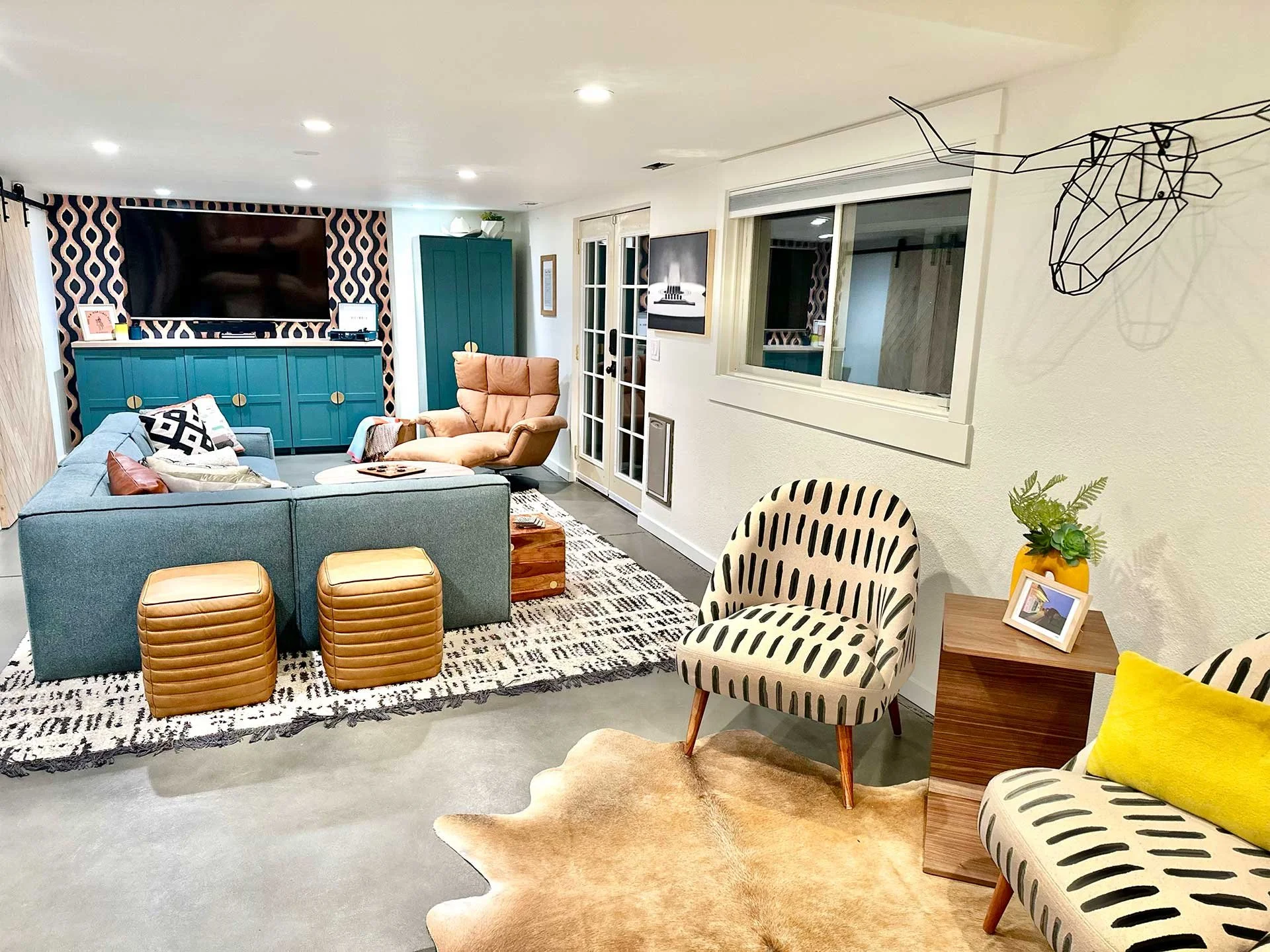 Stylish basement living room with gray sectional, leather lounge chair, patterned accent chairs, layered rugs, and teal built-in media wall with bold wallpaper backdrop.