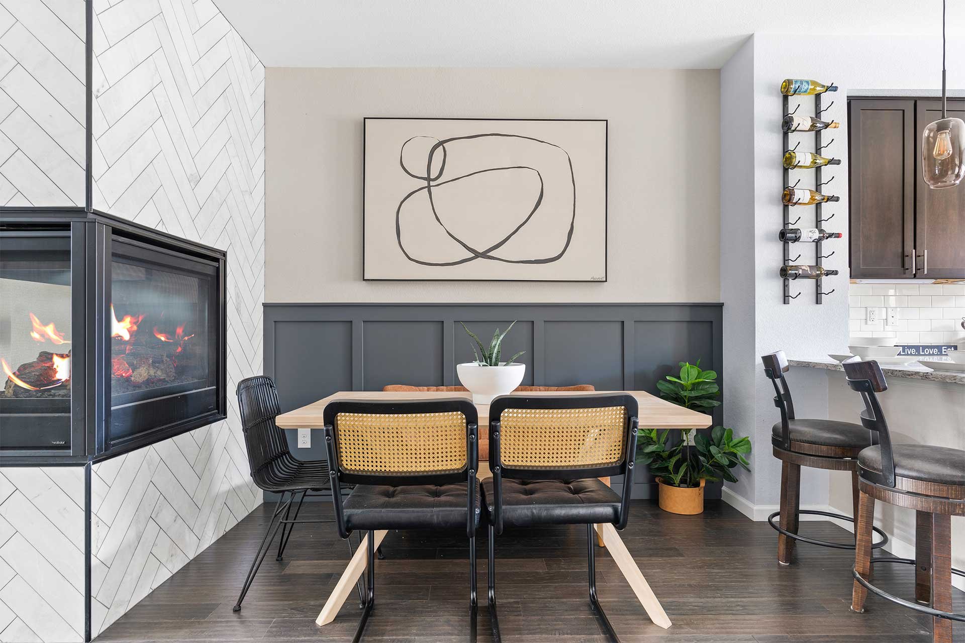 Modern dining space design with herringbone tile wall, abstract black art, dark gray wainscoting, rattan chairs, plants, and fireplace.