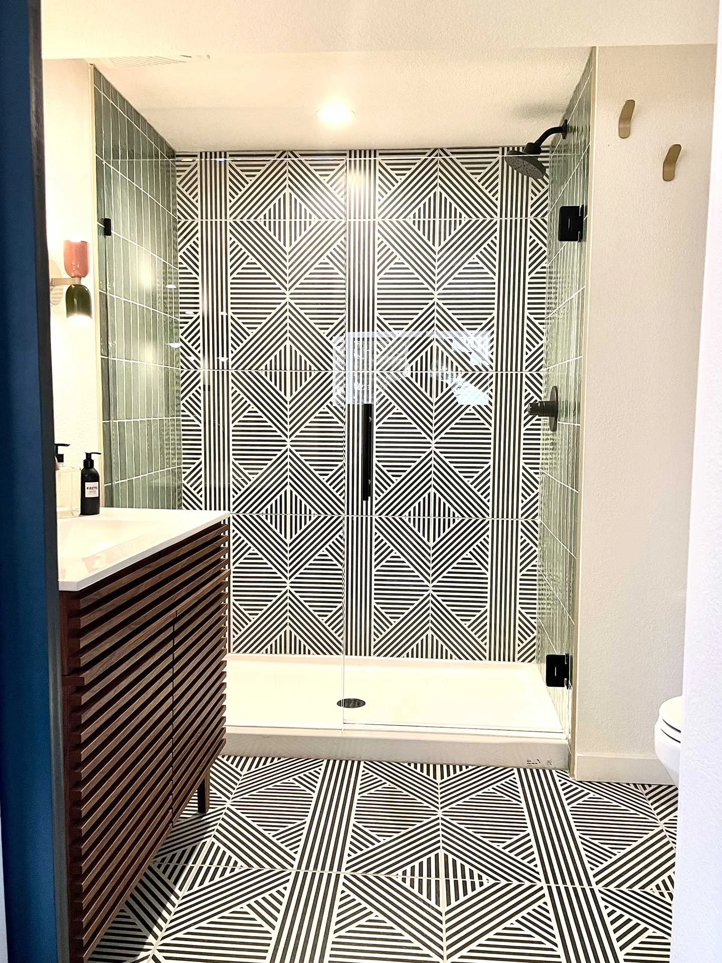 Modern bathroom with glass shower, bold black-and-white geometric tile, green vertical wall tile, and wood slat vanity with clean white countertop.