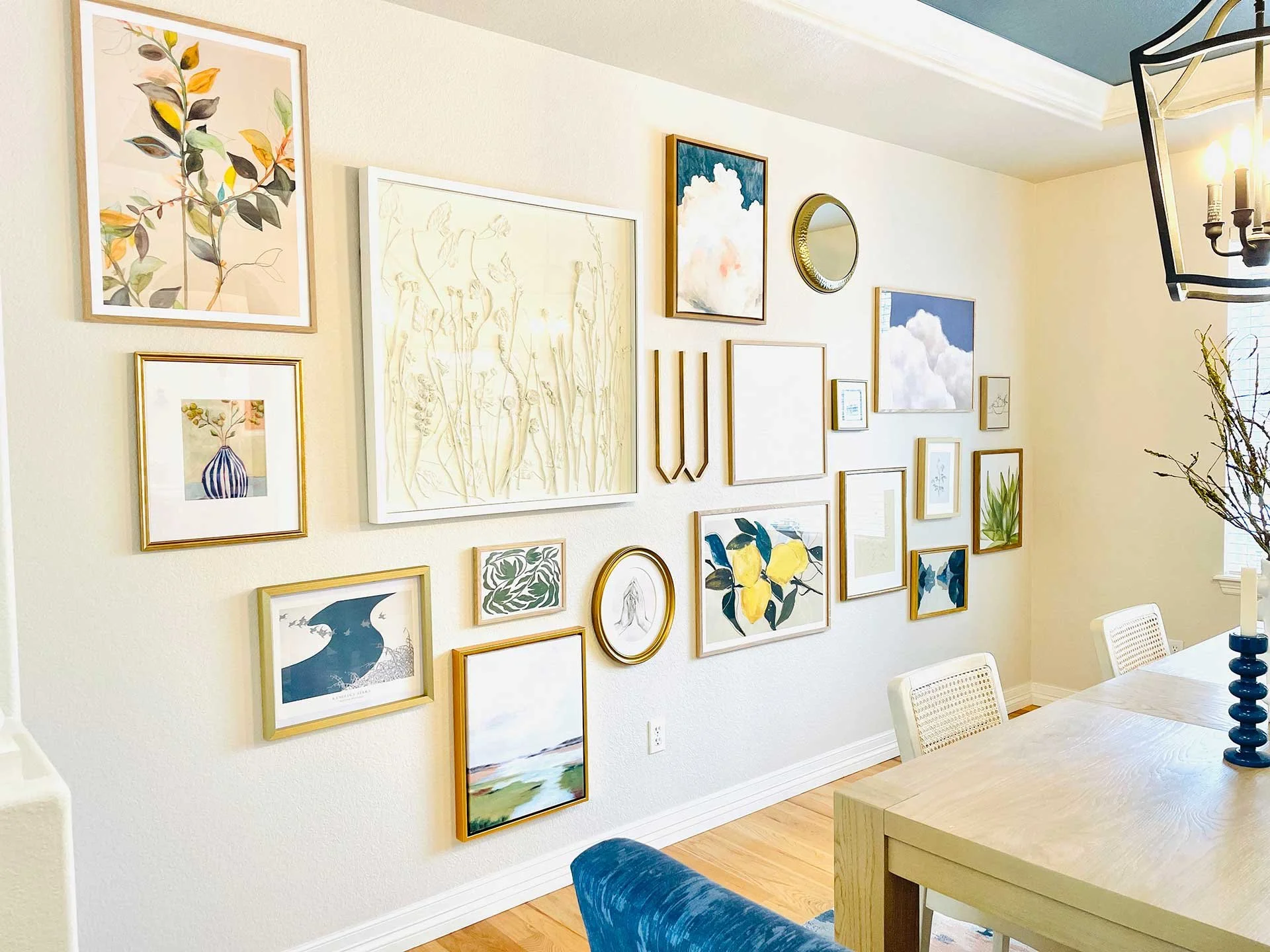Dining room gallery wall with mixed artwork in gold frames, soft neutral palette, and light wood table with woven chairs and styled decor.