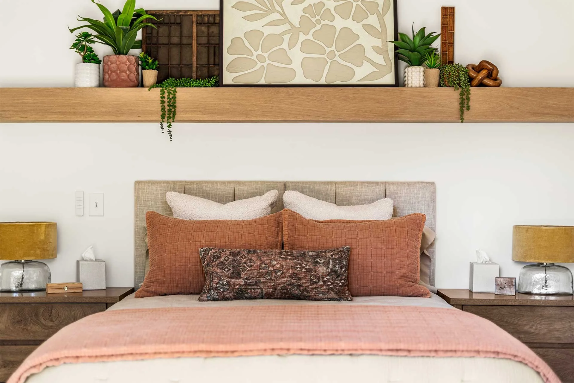 Stylish oak mantel shelf with trailing plants, potted greenery, beige leaf art, wooden decor, and pink vase above a cream headboard