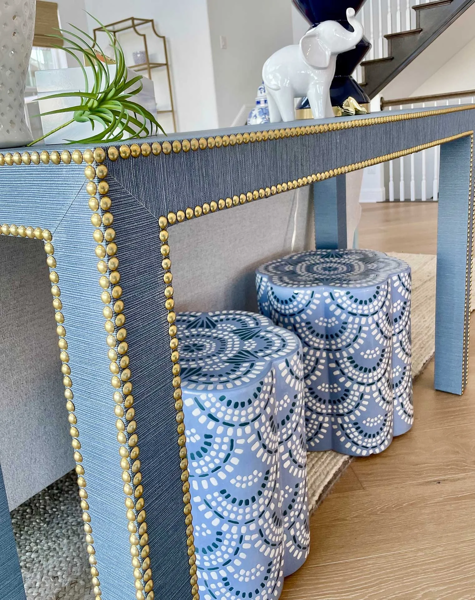 Close-up of blue grasscloth console table with gold nailhead trim, styled with ceramic stools, decorative objects, and greenery in a bright entry space.
