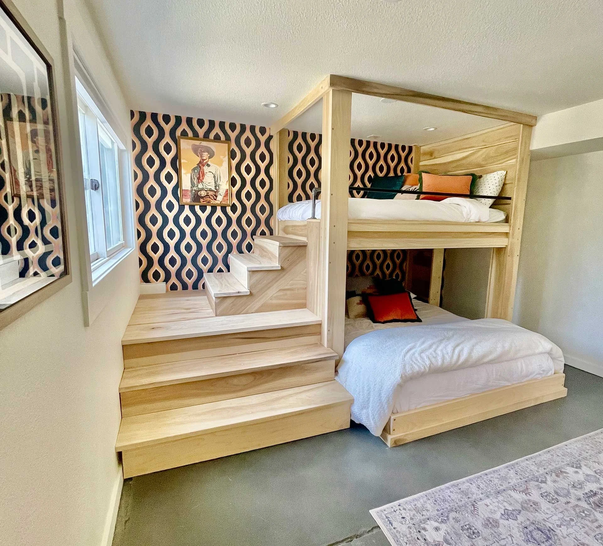 Custom wood bunk beds with built-in stairs and railing, set against bold patterned wallpaper, with layered bedding and warm, modern finishes.
