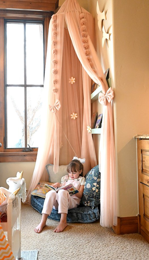 Whimsical play space by full service interior design firm: girl in pink pajamas on beanbag, pink canopy, books, and wooden nook.
