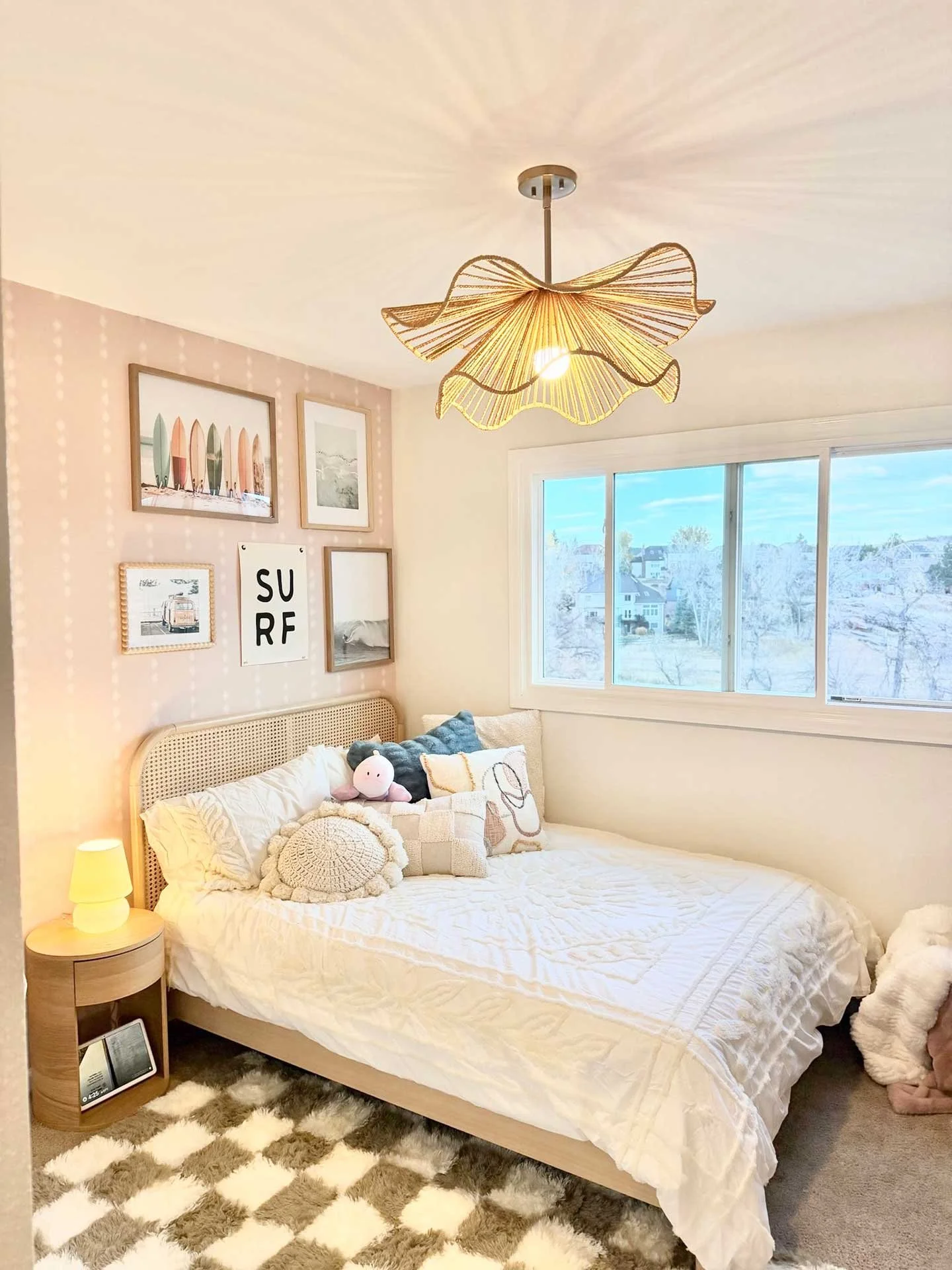 Surf-themed teen bedroom with cane daybed, blush patterned accent wall with surf-inspired gallery art, woven pendant light, and cozy layered bedding beside a large window.