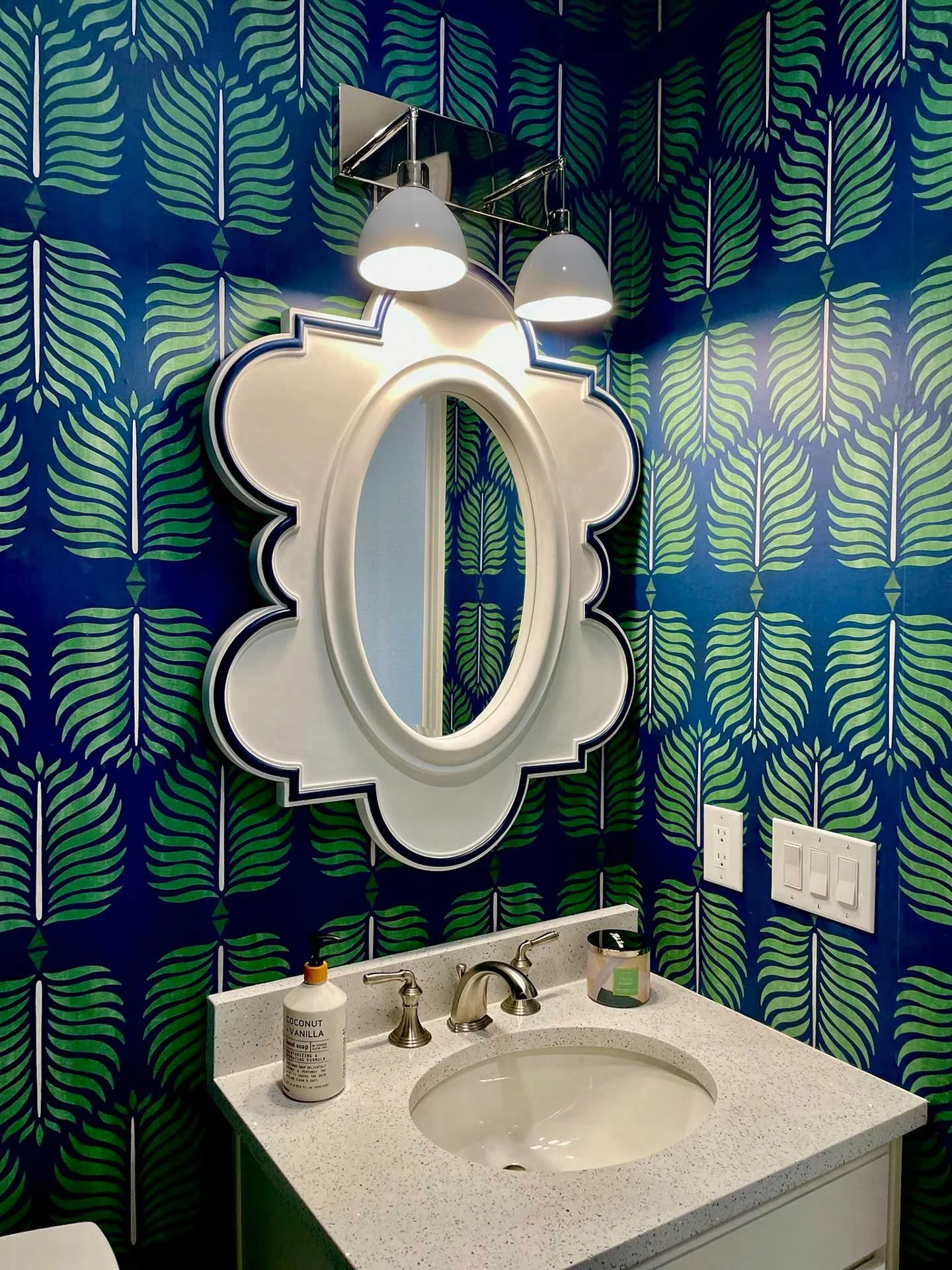Close-up of powder room vanity with white scalloped mirror, double sconce lighting, and bold blue and green leaf-pattern wallpaper.