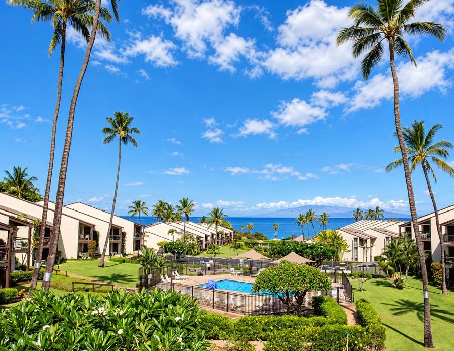 The beautiful grounds of Hale Kamaole, Palm trees, a swimming pool, green lawn, and ocean view under blue sky.