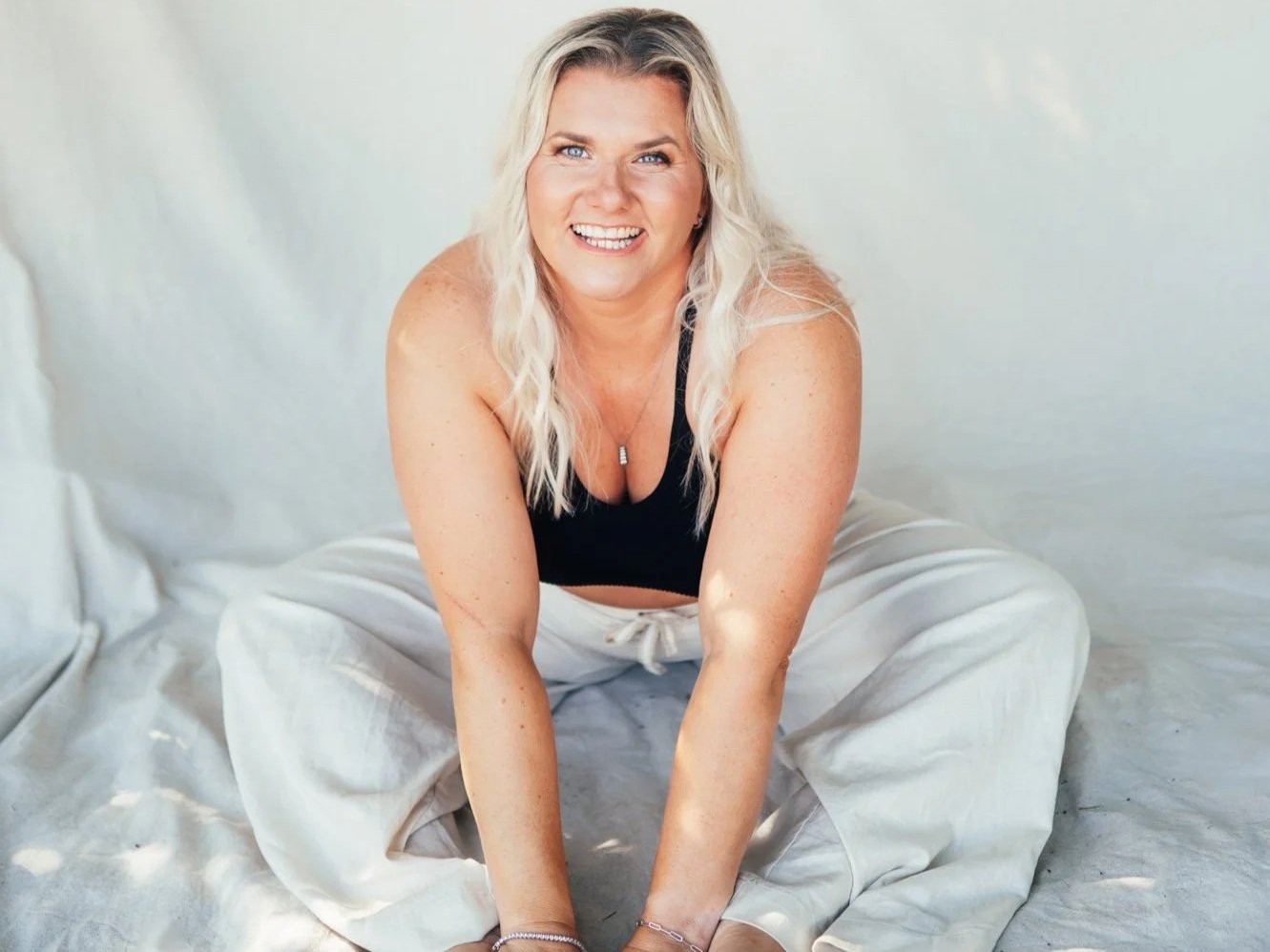 A woman with long blonde hair, wearing a black tank top and white pants, smiling and sitting on the floor in a bright room with a plain white background.
