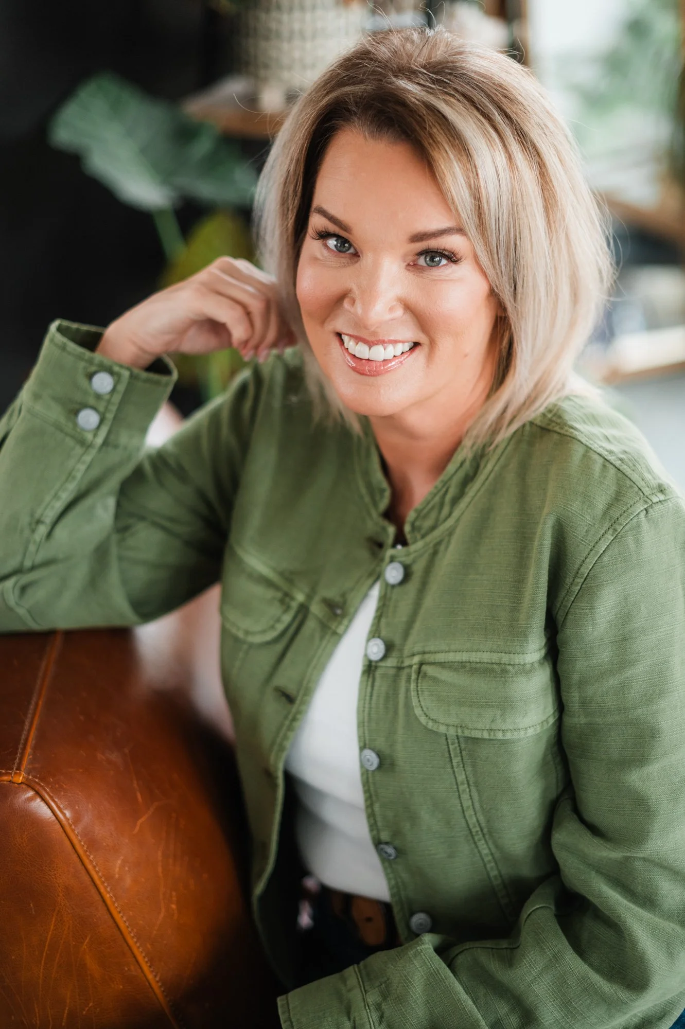 A woman with blonde hair wearing a green jacket, sitting on a leather couch, smiling at the camera.