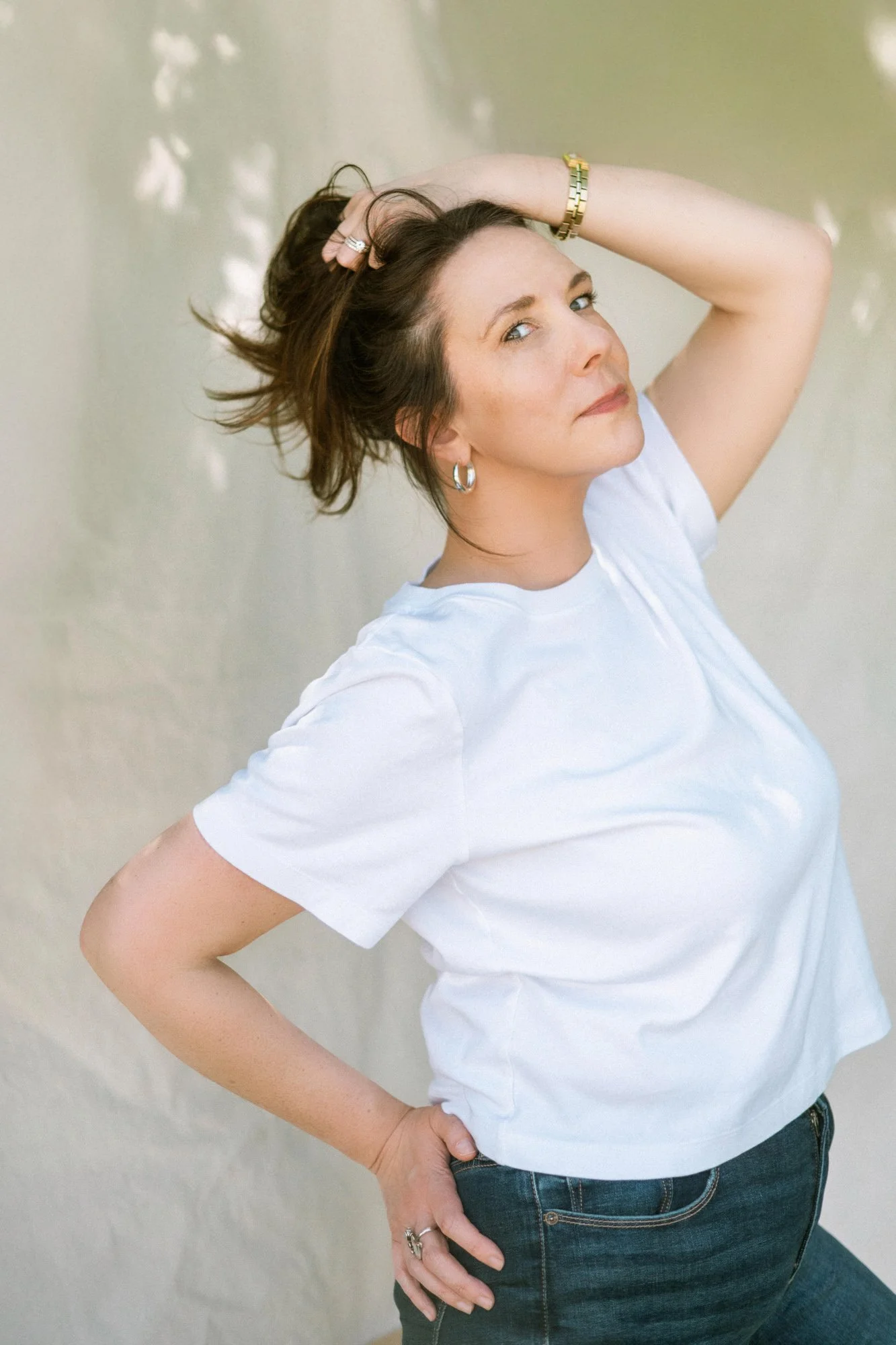 A woman with shoulder-length brown hair, wearing a white t-shirt and dark jeans, poses with one hand on her hip and the other behind her head, looking at the camera with a confident expression against a plain, lightly textured wall.