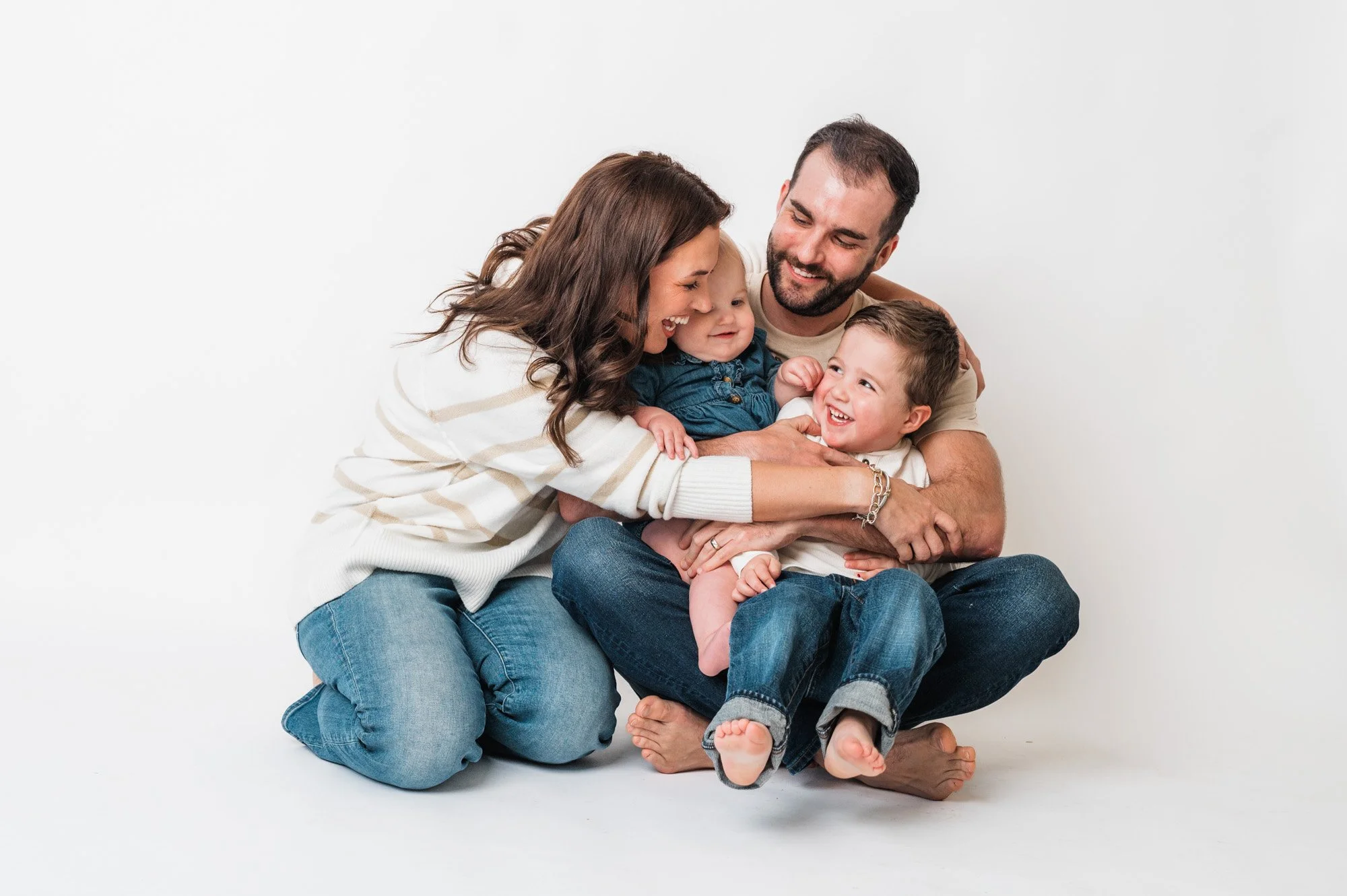 A smiling family of four, with two children, sitting and playing against a plain white background.
