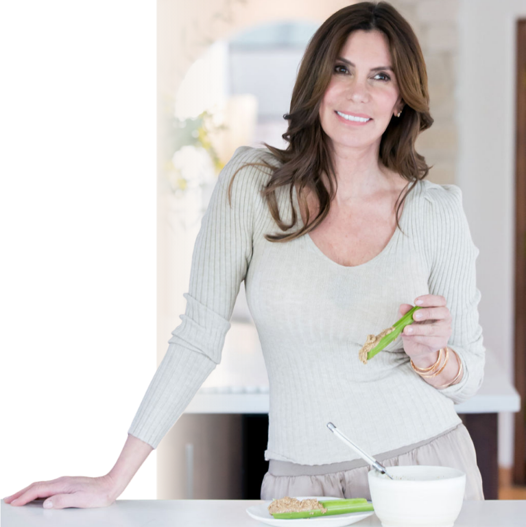 Lizanne Falsetto leaning on her kitchen counter, holding a celery stalk with nut butter