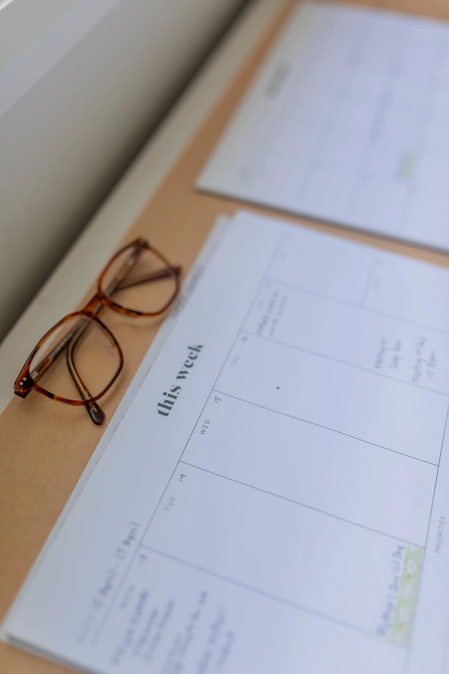 A calendar page on a desk with a pair of tortoiseshell glasses placed on it, and some papers behind. The calendar shows part of the week and some handwritten notes.