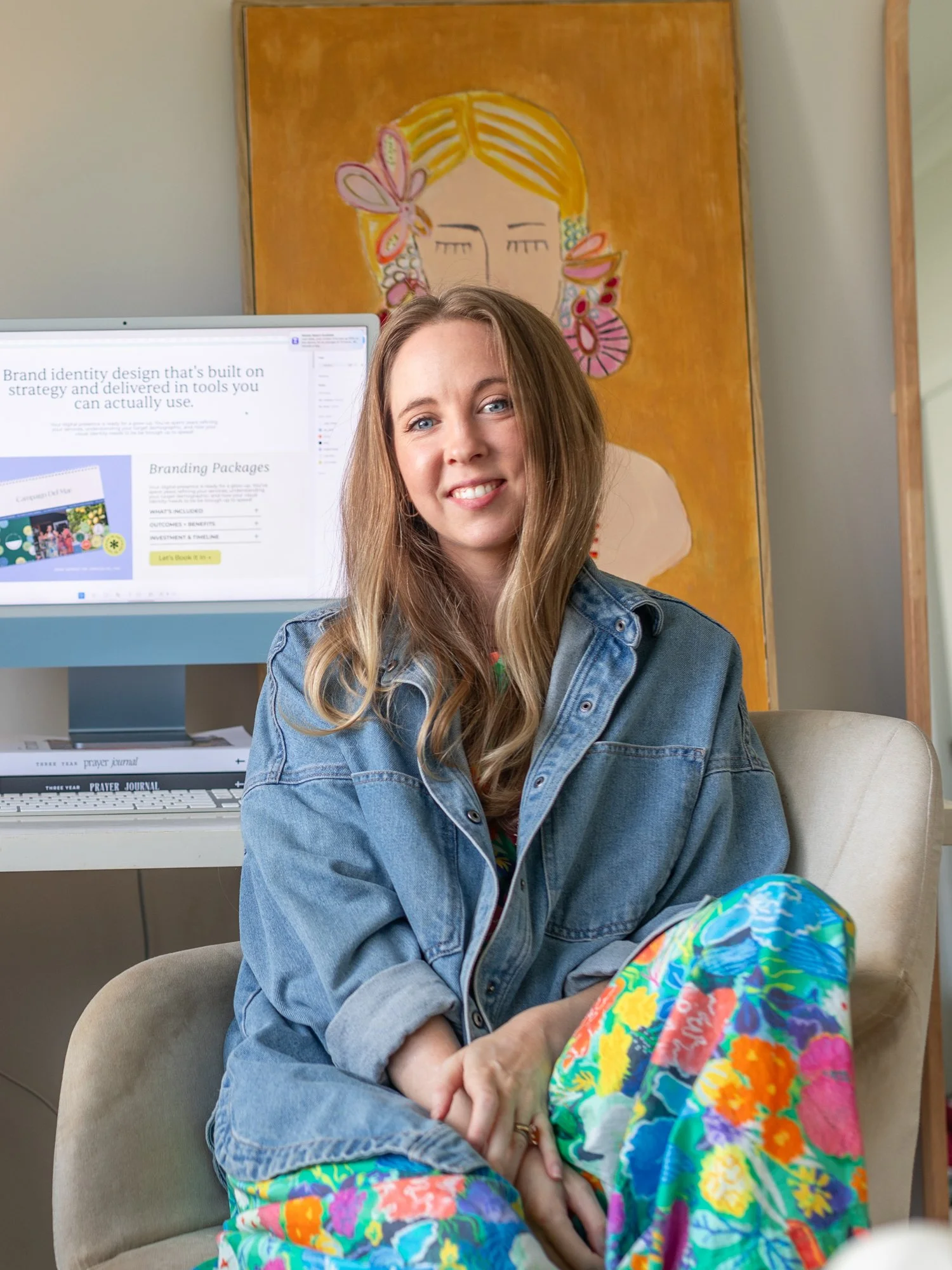 A smiling young woman with long light brown hair sitting in a beige armchair, wearing a denim jacket and colorful floral pants, in front of a computer monitor displaying a website, with a colorful abstract portrait of a woman's face with closed eyes and floral earrings hanging behind her.