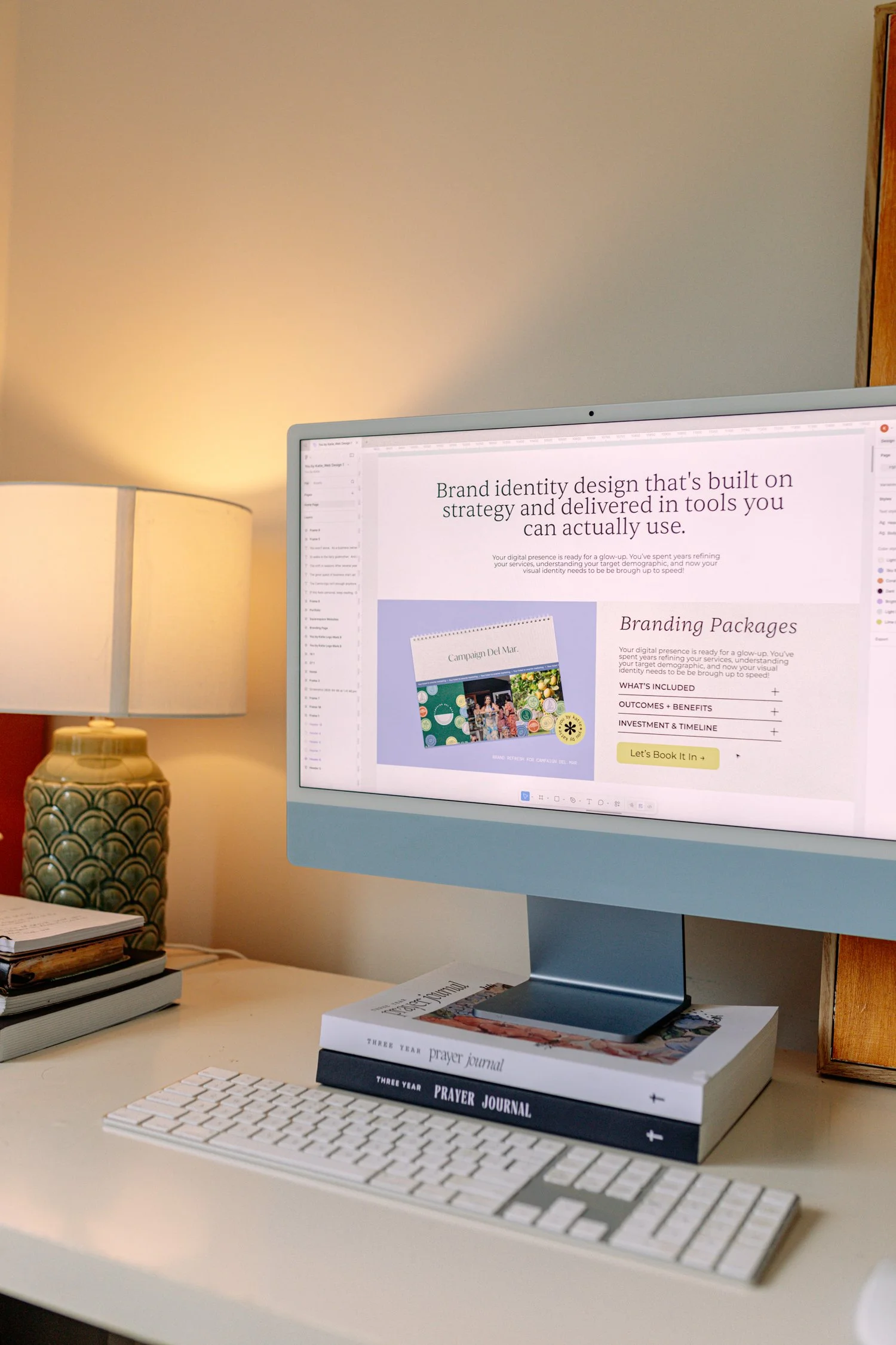 Computer monitor displaying a webpage about brand identity design, with a lamp, notebooks, and prayer journals on the desk.