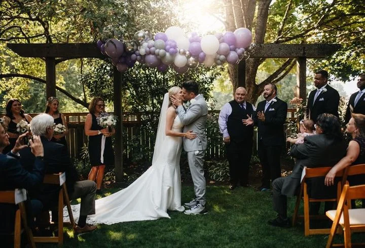 wedding-couple-ceremony-kiss-under-purple-white-balloon-garland-outdoors-yelm-washington.jpg