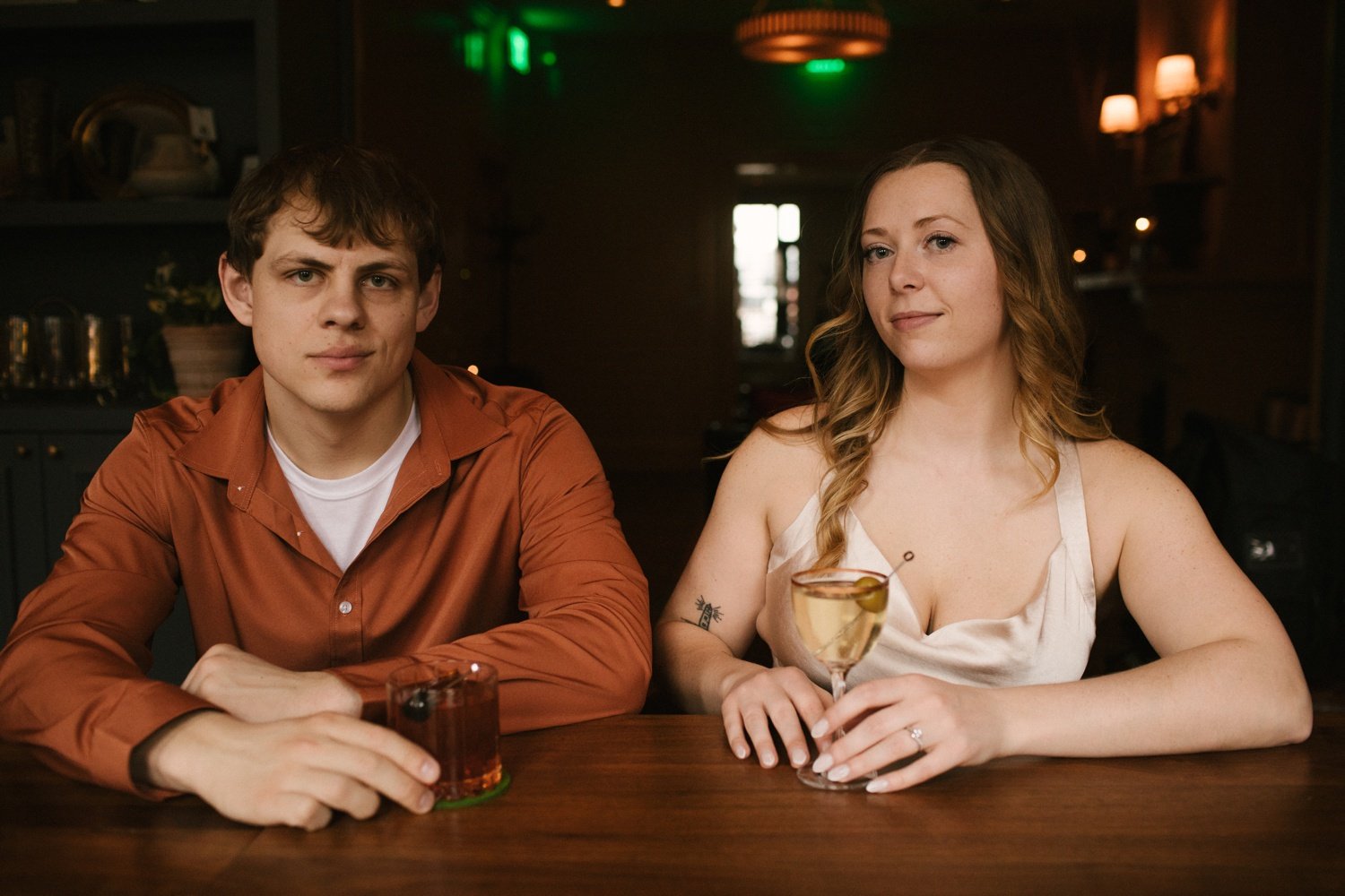 man and woman sit at table with fancy cocktails in dress clothes for engagement session at the daisy lounge in downtown stevens point wisconsin
