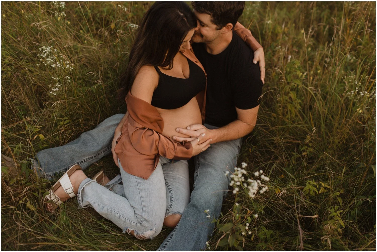 pregnant woman sits in husbands lap with hands on belly in a field of long grass and white flowers for maternity session in wisconsin rapids
