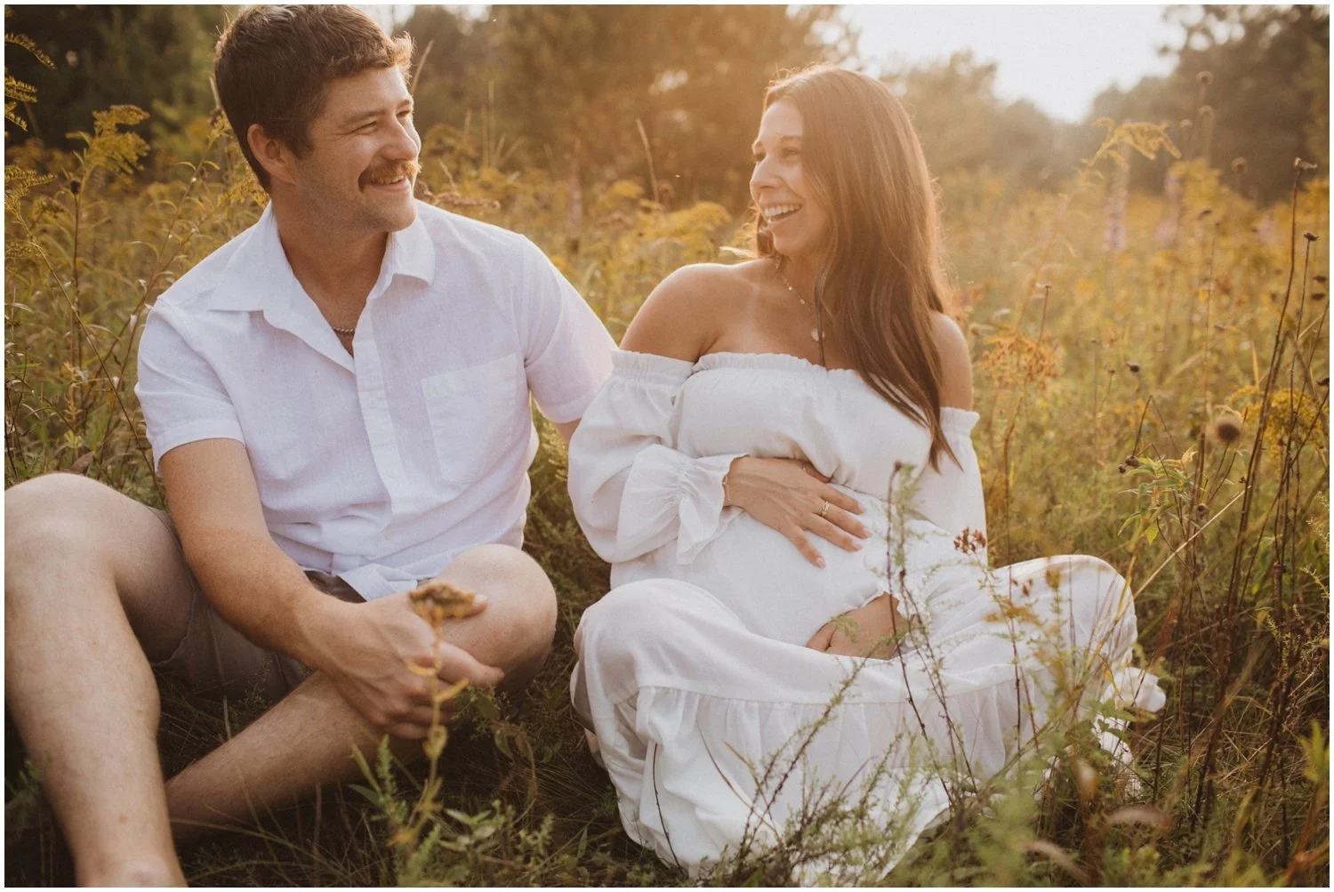 a pregnant woman and her husband wearing white sit in a field of wild flowers for maternity session in wisconsin rapids