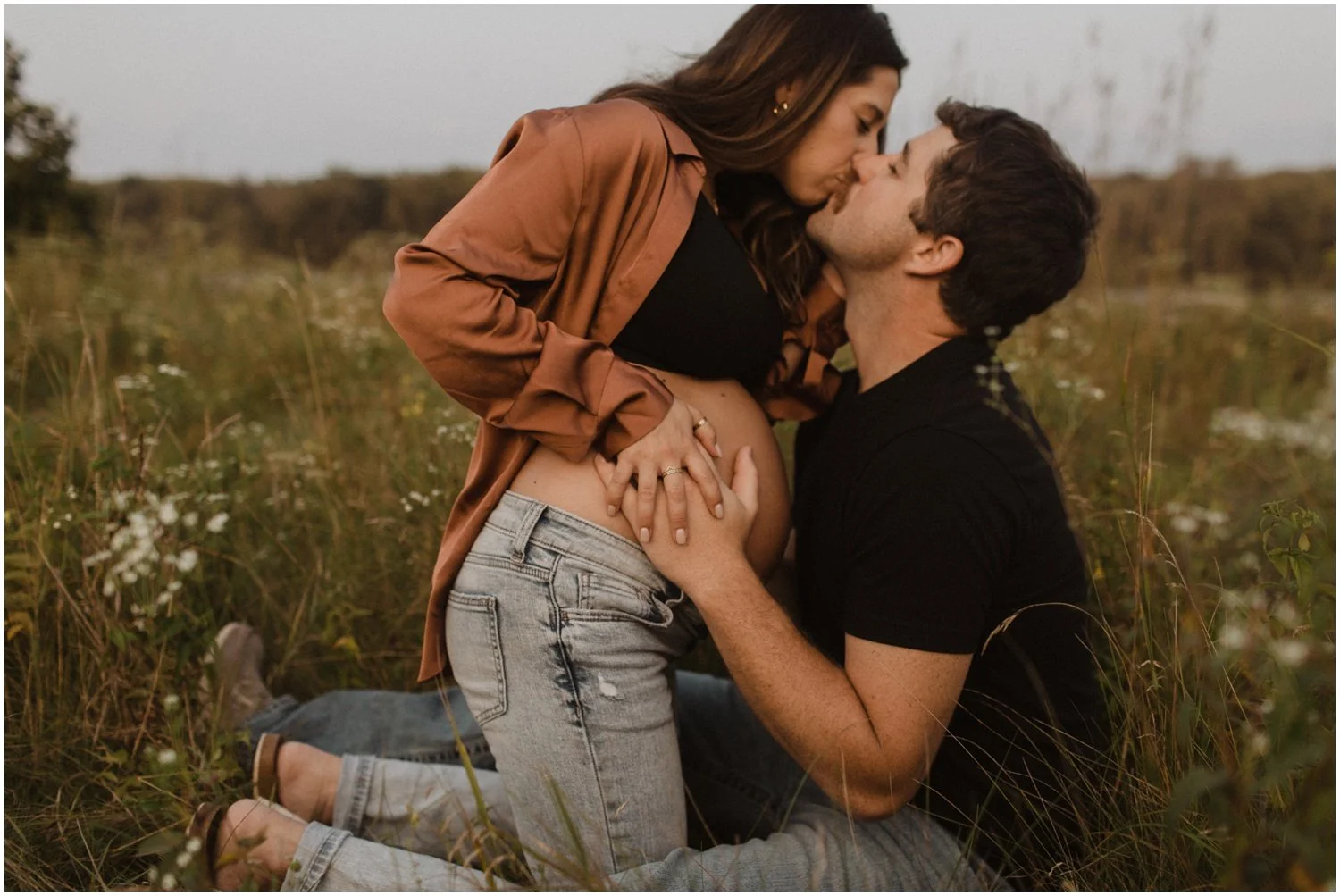 pregnant woman kneels in front of husband while they kiss and have hands on belly for maternity session in a field of long grass and white flowers wisconsin rapids