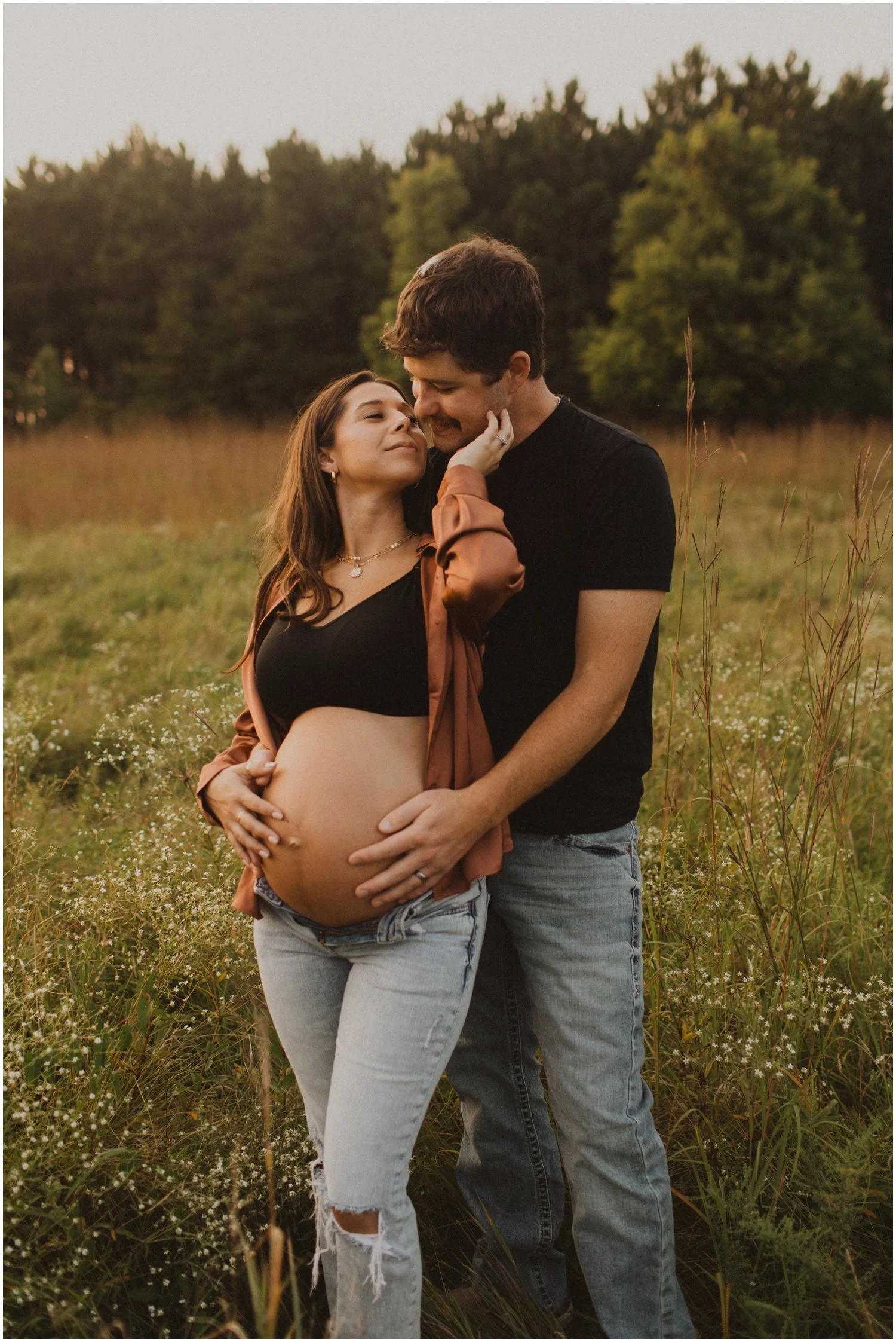 pregnant woman standing in front her husband with hands on belly for maternity session in a field of long grass in wisconsin rapids