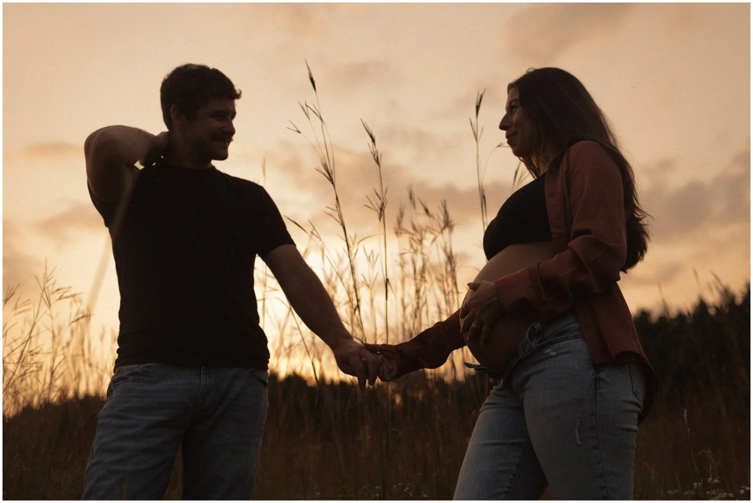 silhouette of pregnant woman and her husband holding hands in a field with long grass in wisconsin rapids