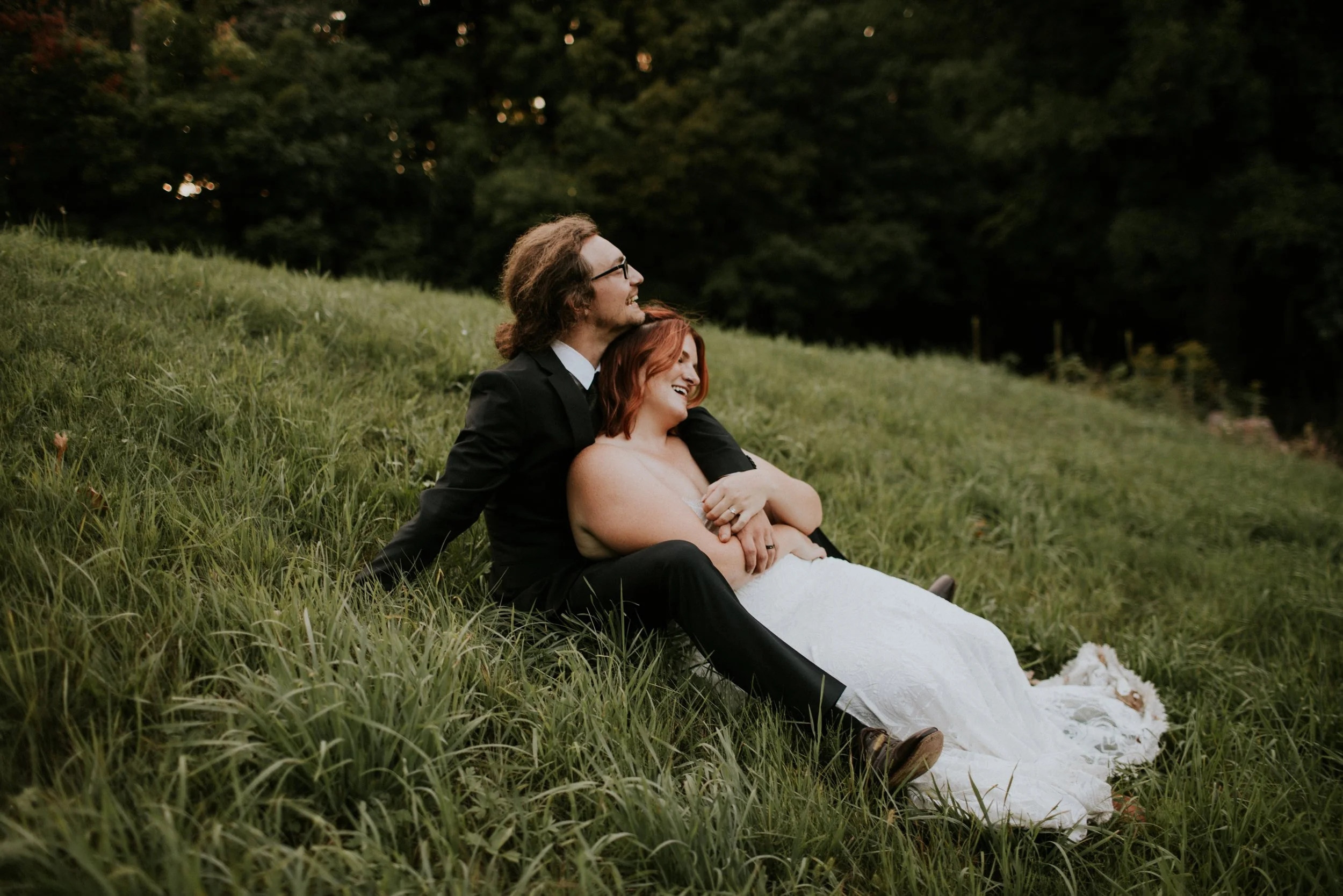 a bride and groom sitting on a grassy hill leaning into each other and laughing at powers bluff park in marshfield wisconsin