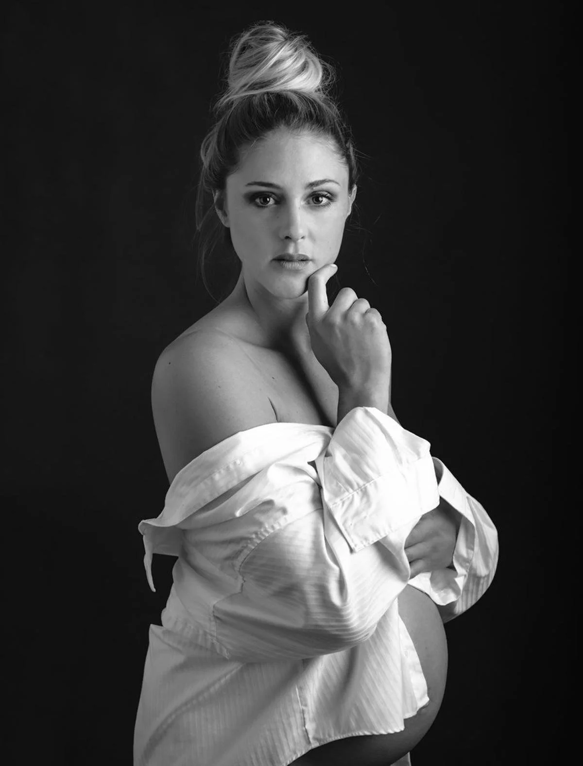 Black and white portrait of a pregnant woman with her shirt off, looking directly into the camera with her right hand near her chin and a thoughtful expression.