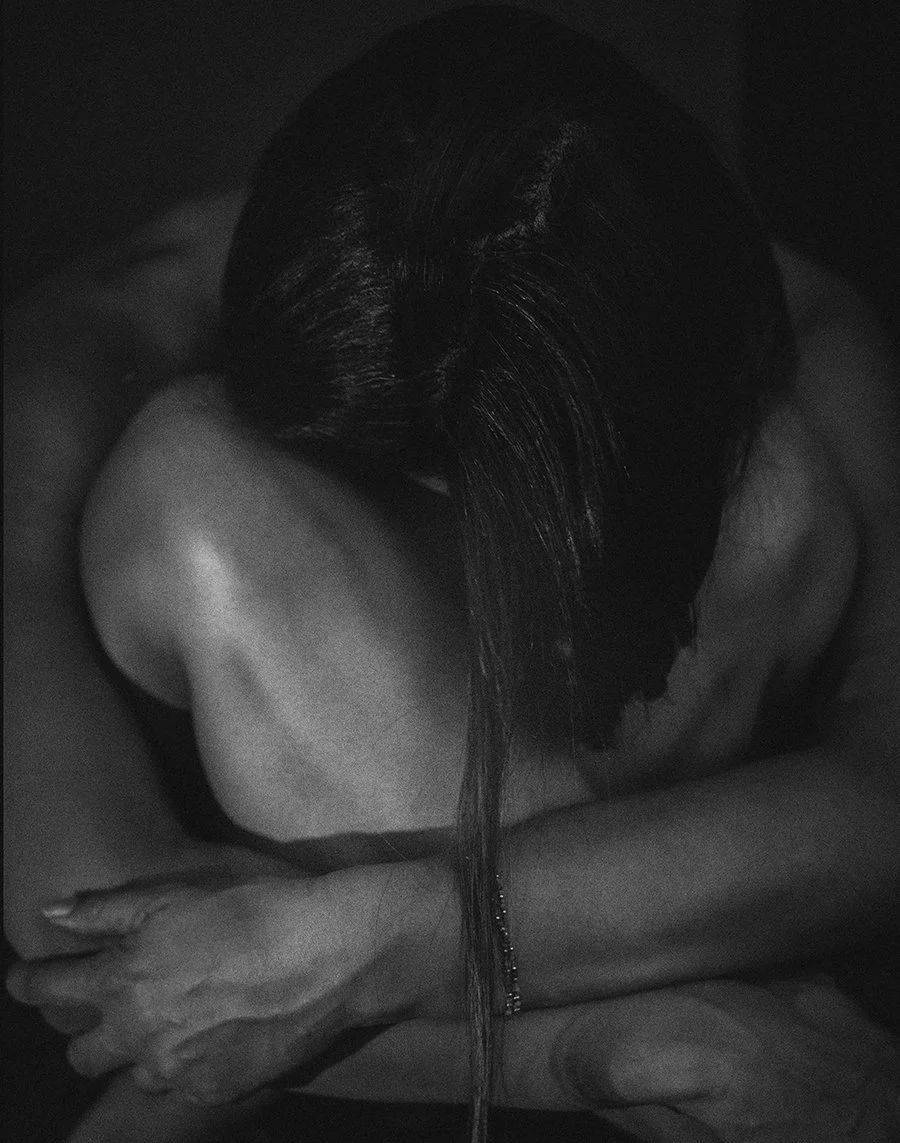 A person with dark hair sitting on their knees with their arms wrapped around their legs, head bowed down, in a black and white photo.