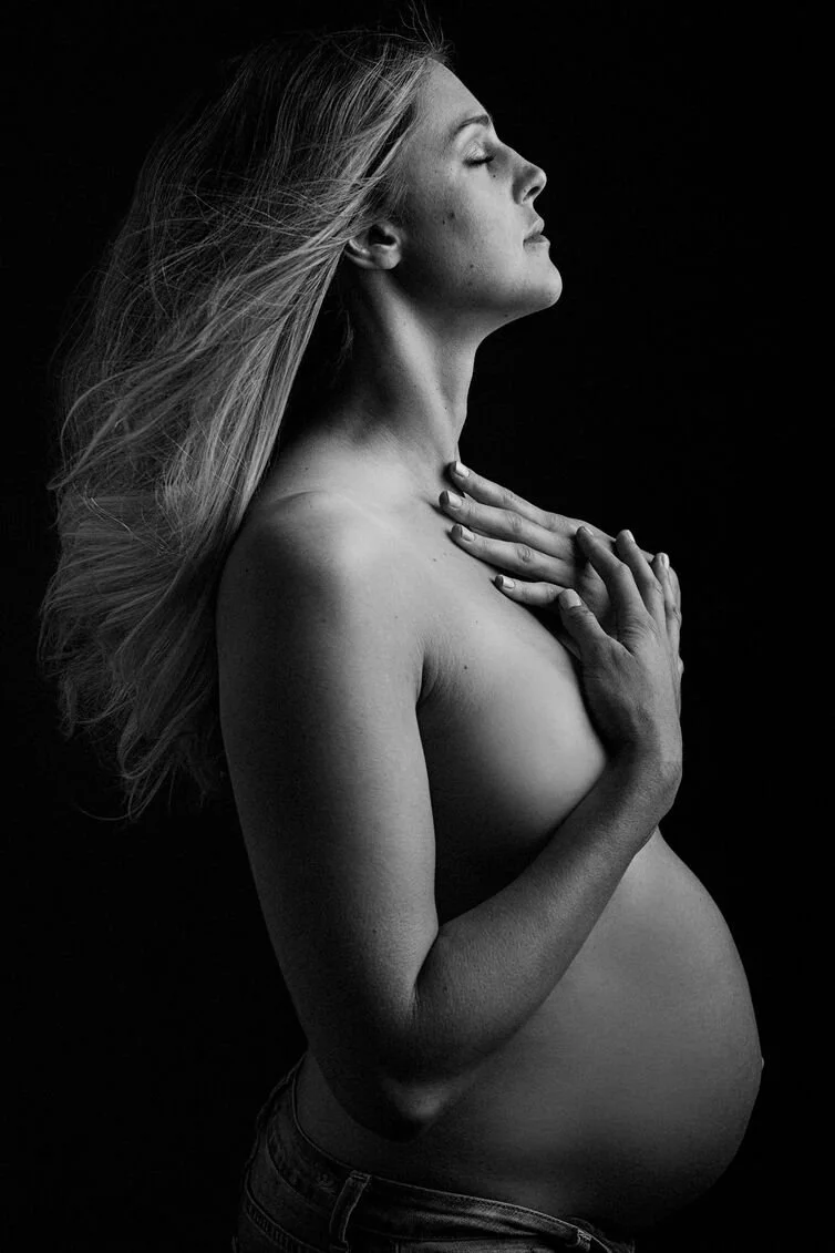 A pregnant woman with long hair, closed eyes, and a serene expression, holding her chest with one hand and resting her other hand on her belly, photographed in black and white against a dark background.