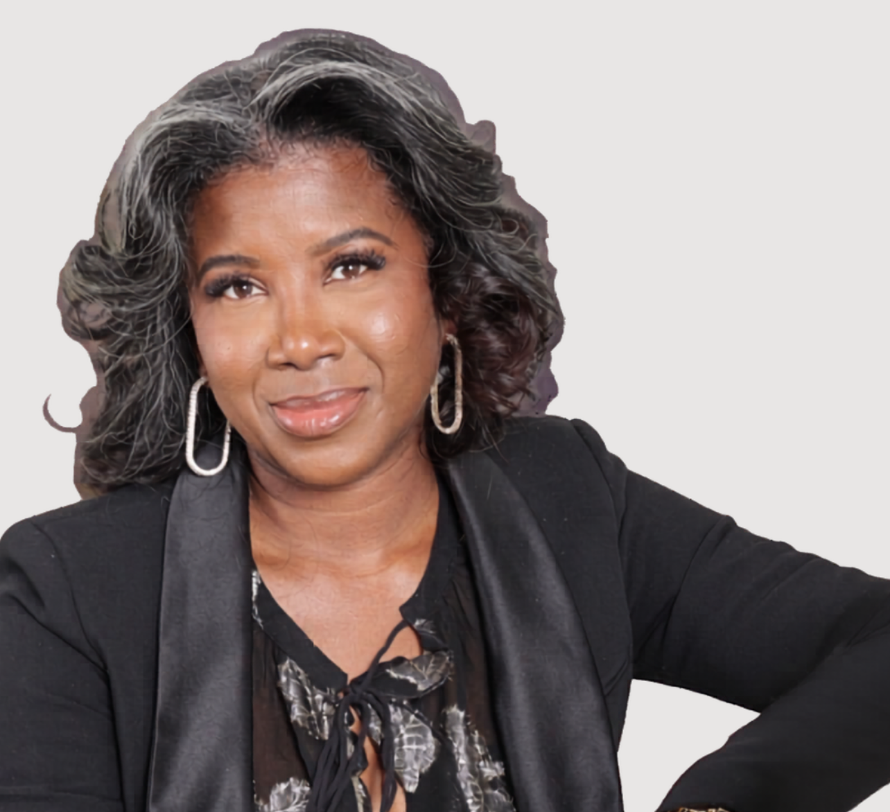 Portrait of a confident Black woman with shoulder-length curly hair, wearing a black blazer, earrings, and a patterned blouse, against a light background.