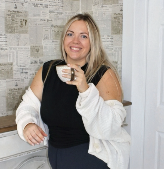 A woman holding a cup of coffee, smiling, with blonde hair, wearing a black top and a white cardigan, standing in front of a wall decorated with newspaper clippings.