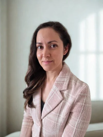 A woman with long dark hair wearing a light pink plaid blazer, sitting indoors near a window with natural light.