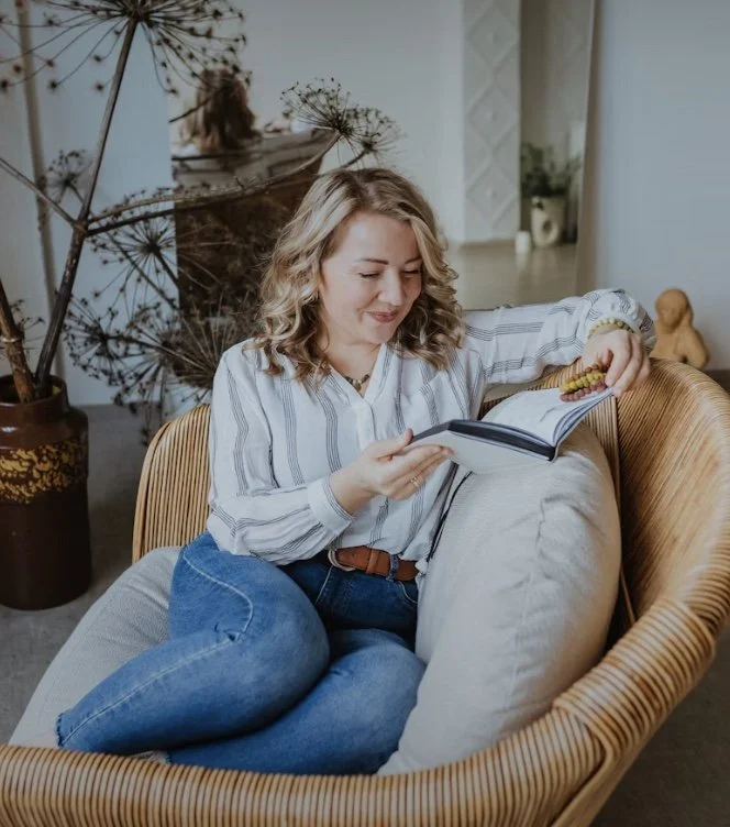 Woman in her 30s sitting comfortably in a cozy chair, smiling while reading a book. Represents self-reflection and emotional wellness in individual therapy.