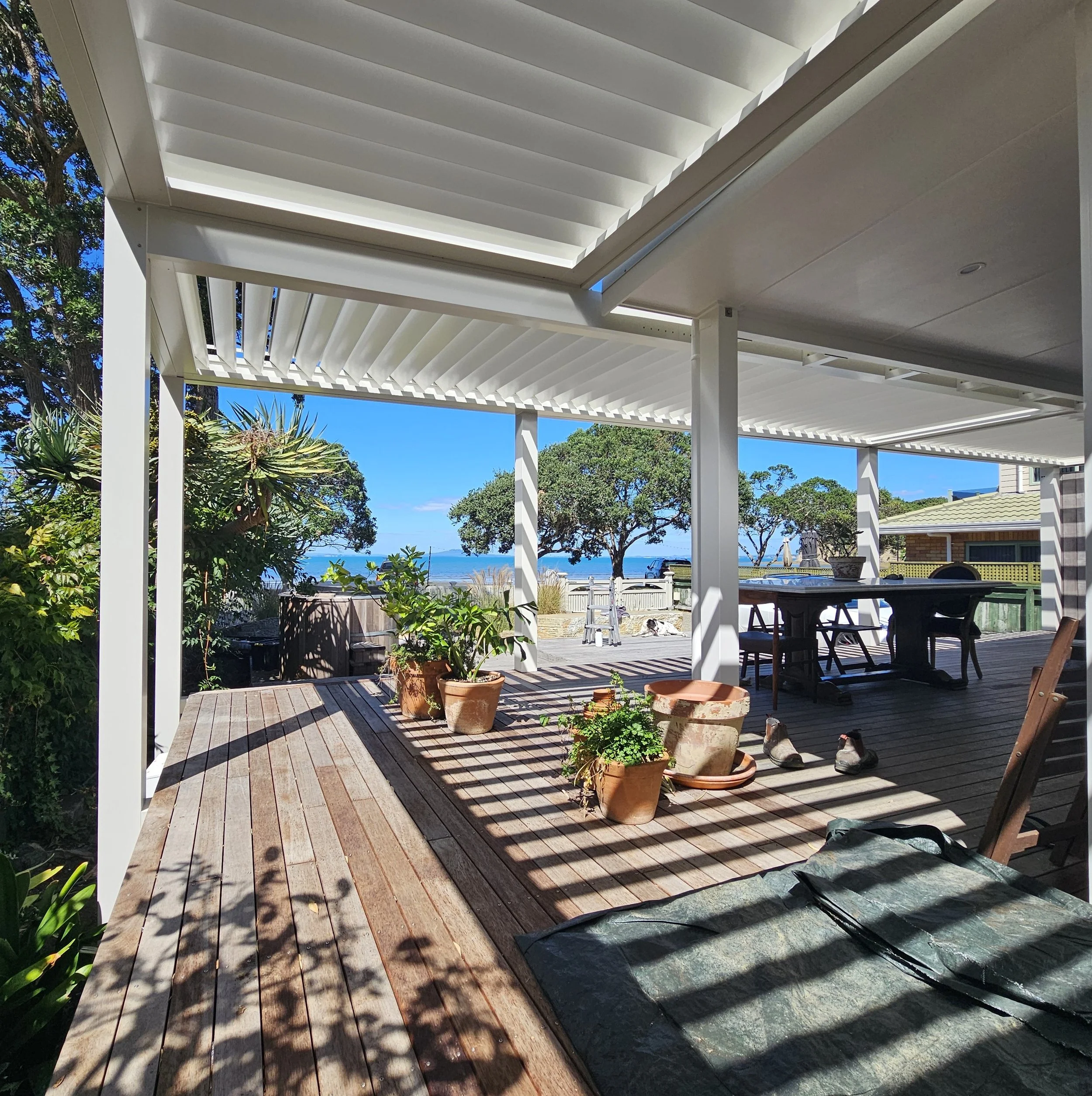 A wooden deck with potted plants and outdoor furniture underneath a white pergola, with trees and the ocean in the background.
