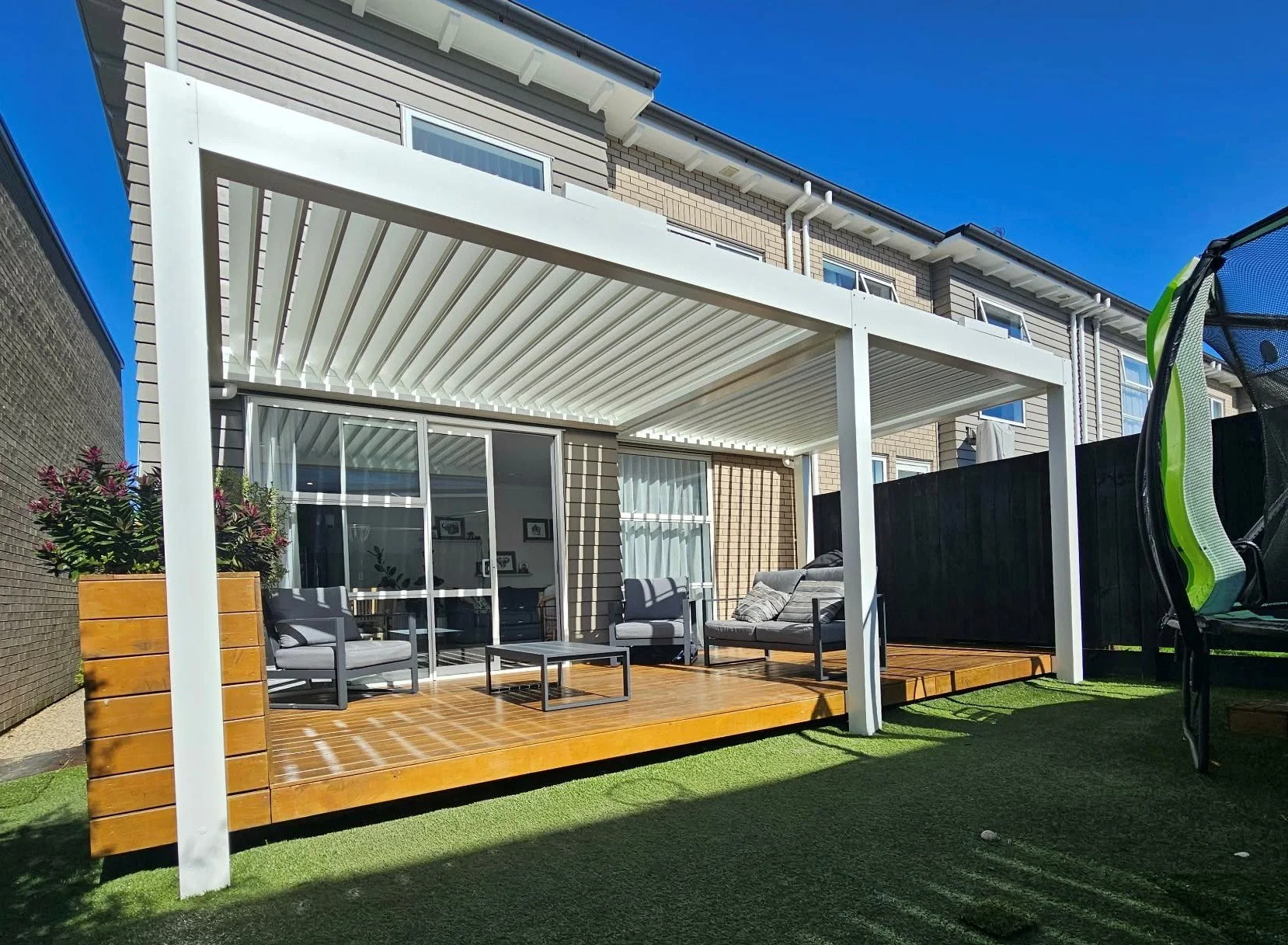 Backyard patio with wooden flooring, outdoor furniture including chairs and a table, potted plants, and a white pergola structure. The patio is adjacent to a house with sliding glass doors, and there is a black fence on the right. The background feat