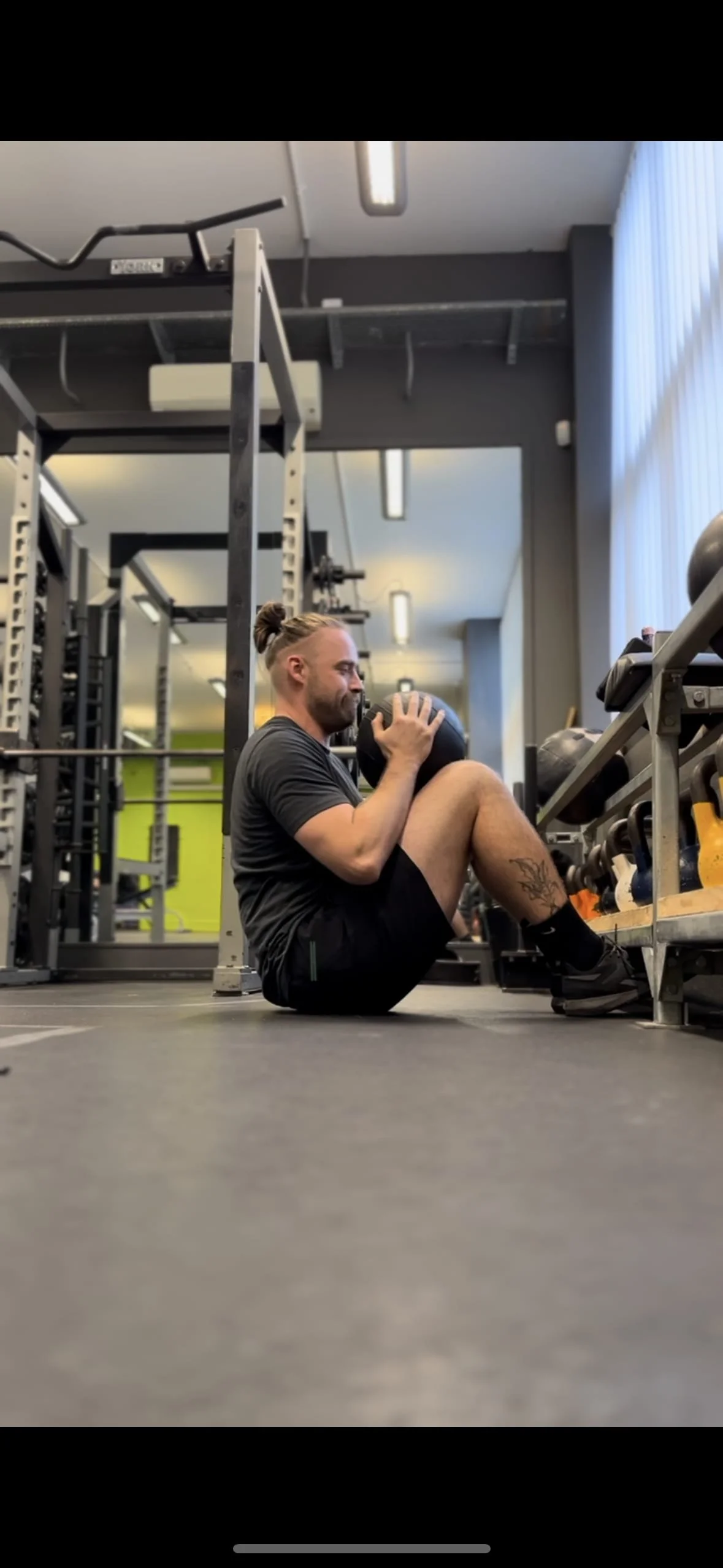 A man with a ponytail and tattoos is sitting on the gym floor, holding a medicine ball close to his chest, in a gym with workout equipment around him. Best personal trainer in Oxford. Best personal trainer near me.