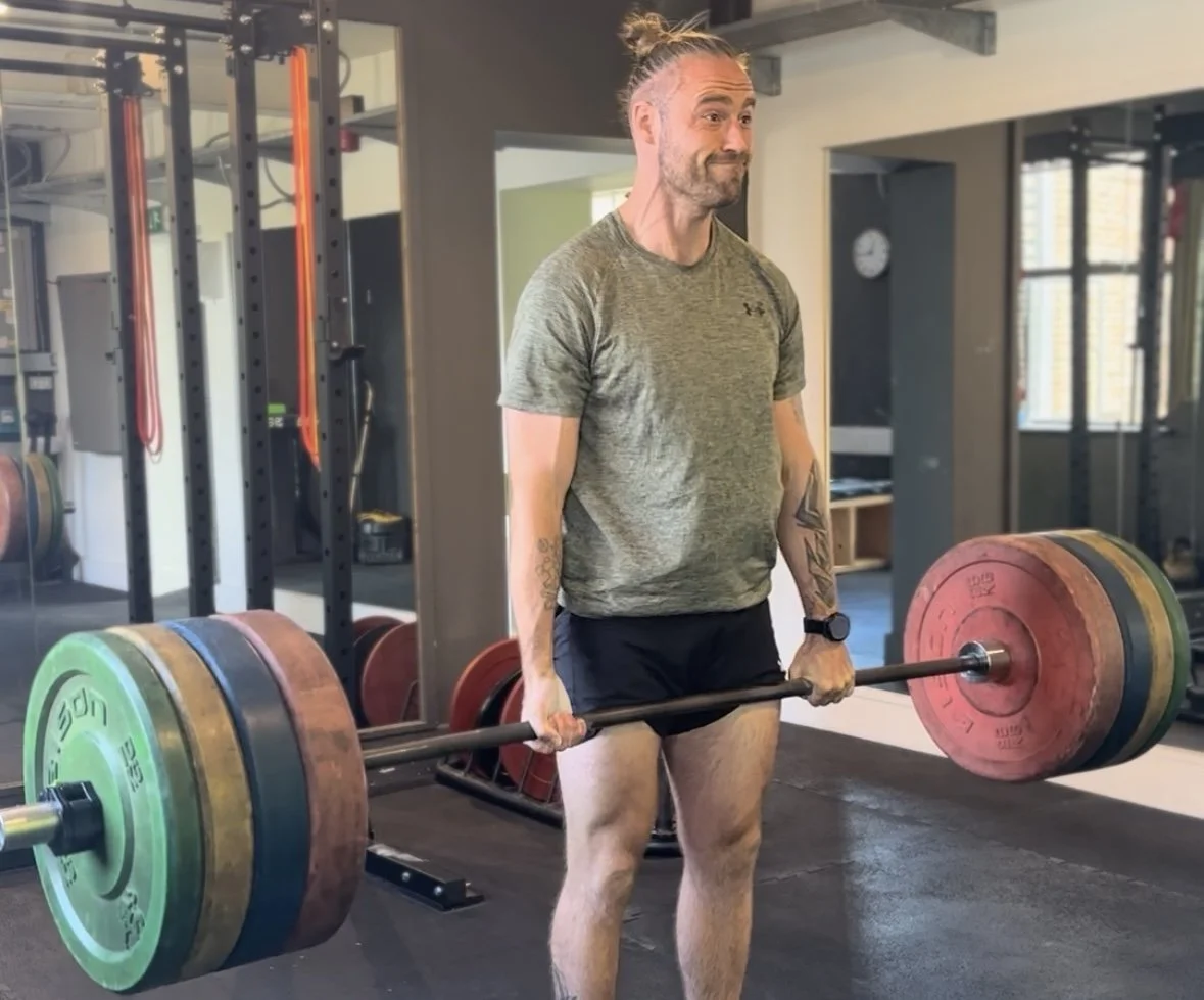 A man with a bun hairstyle, beard, wearing a green athletic shirt and black shorts, is lifting a heavy barbell with colorful weights in a gym. Best personal trainer in Oxford. Best personal trainer near me.