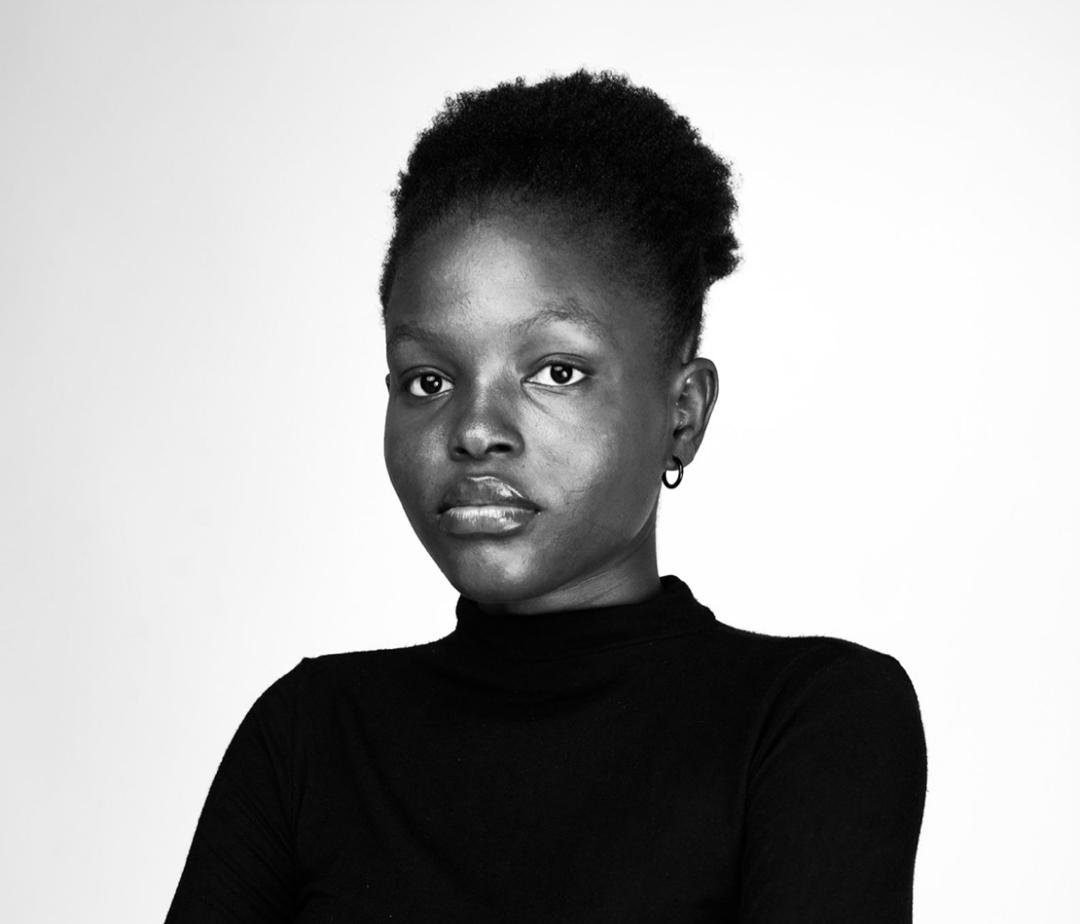 Black and white portrait of a woman with shoulder-length dreadlocks and wearing a black turtleneck and a chain necklace. She is sitting outdoors with a metal fence in the background.