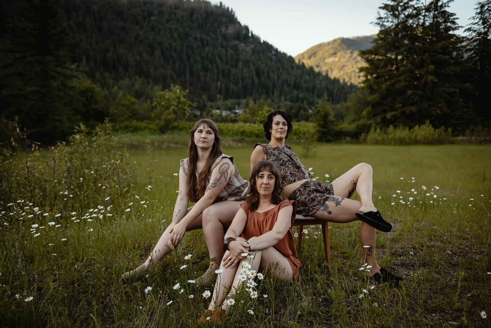 Three women sitting outdoors on a grassy field with daisies, mountain scenery, and trees in the background wearing matching Diane tops and mini bloomers.